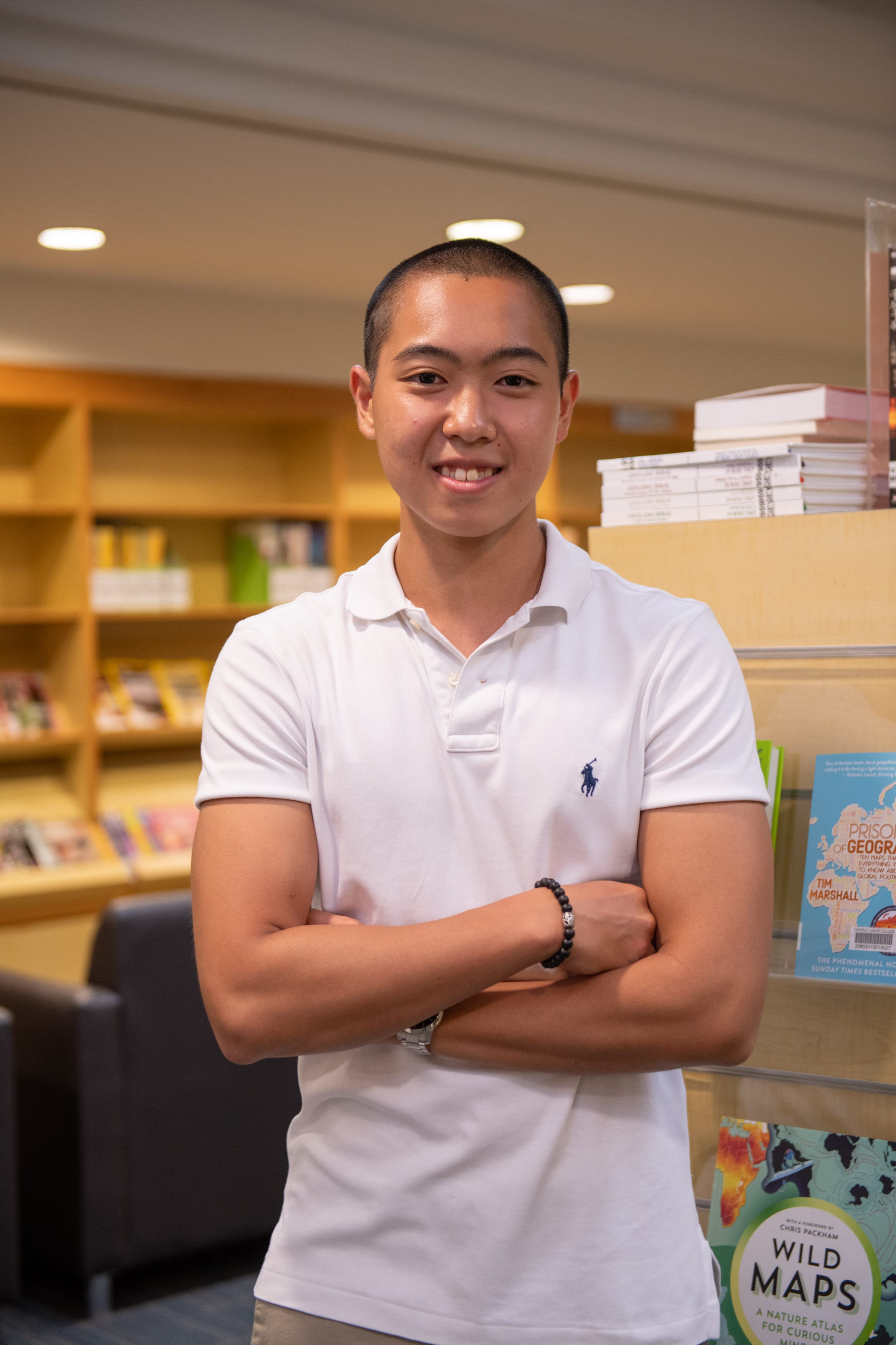 Young man in a white polo shirt smiling with arms crossed in a library with bookshelves behind him.