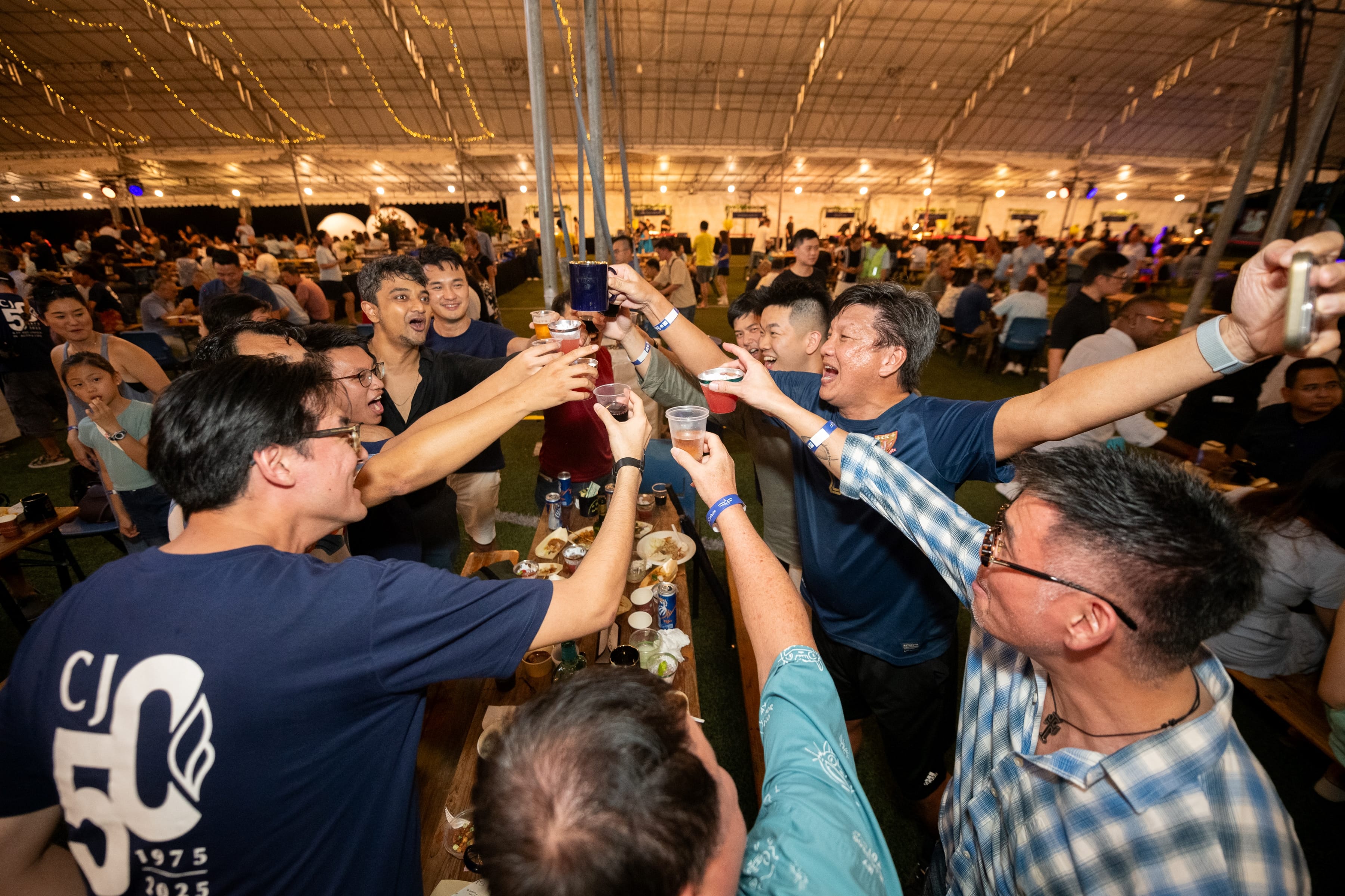 Group of friends at a crowded tent event raising cups to toast over a long table with food and drinks.