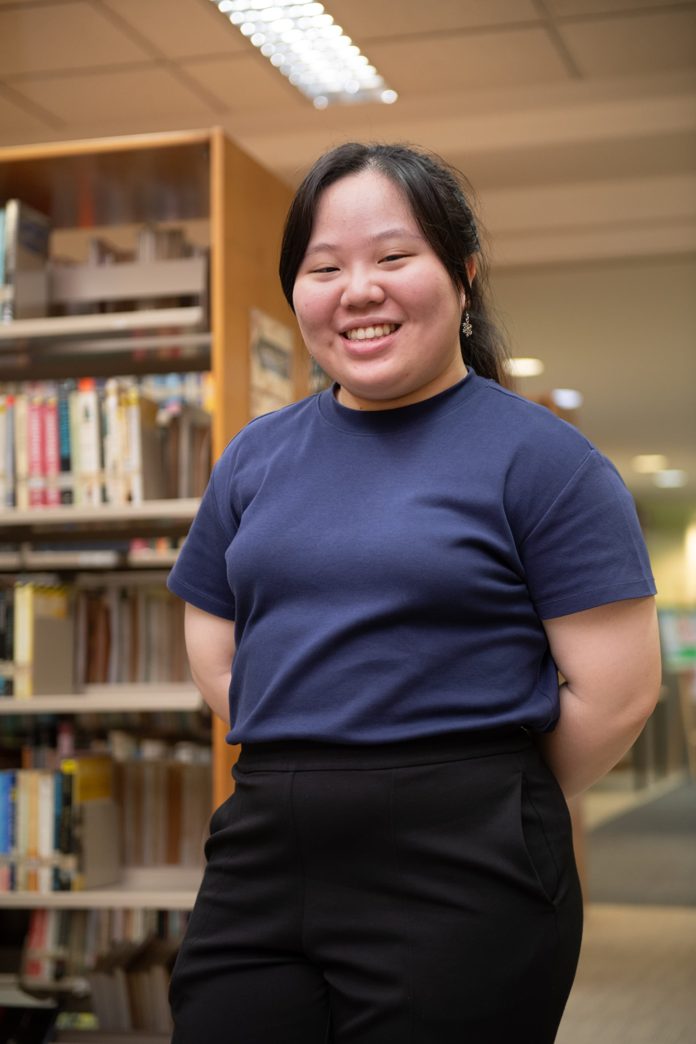 A smiling young woman in a navy blue shirt and black pants stands in front of library bookshelves.
