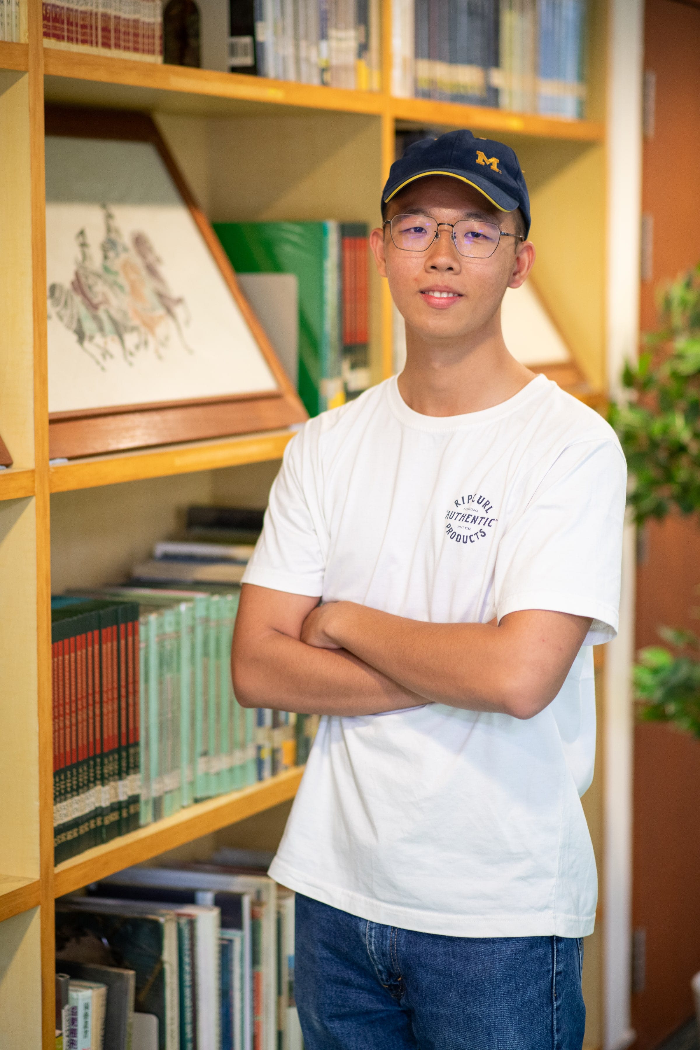 Young man with glasses and a navy cap stands arms crossed in front of wooden bookshelves filled with books.