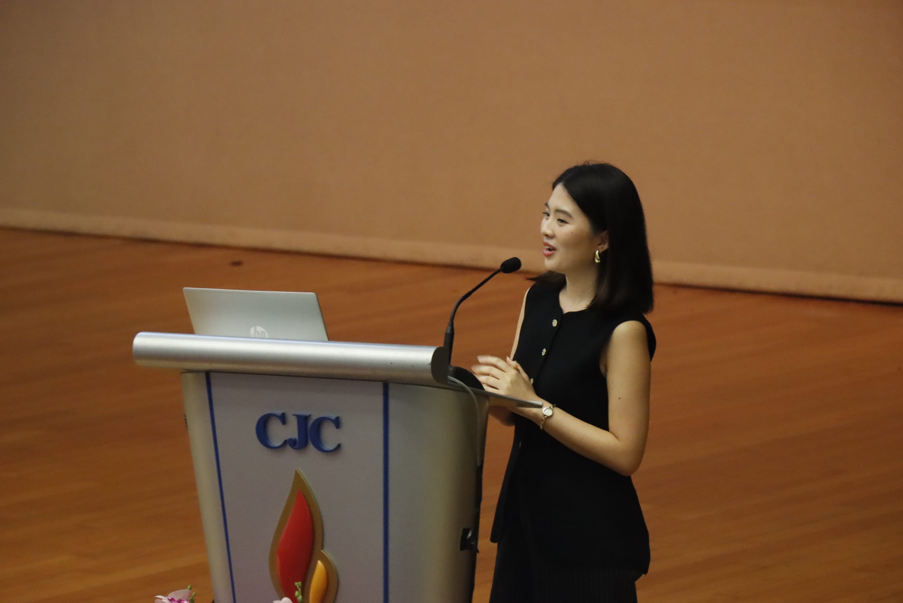 Woman in black dress speaking at a CJC podium with a laptop and microphone on a wooden stage.
