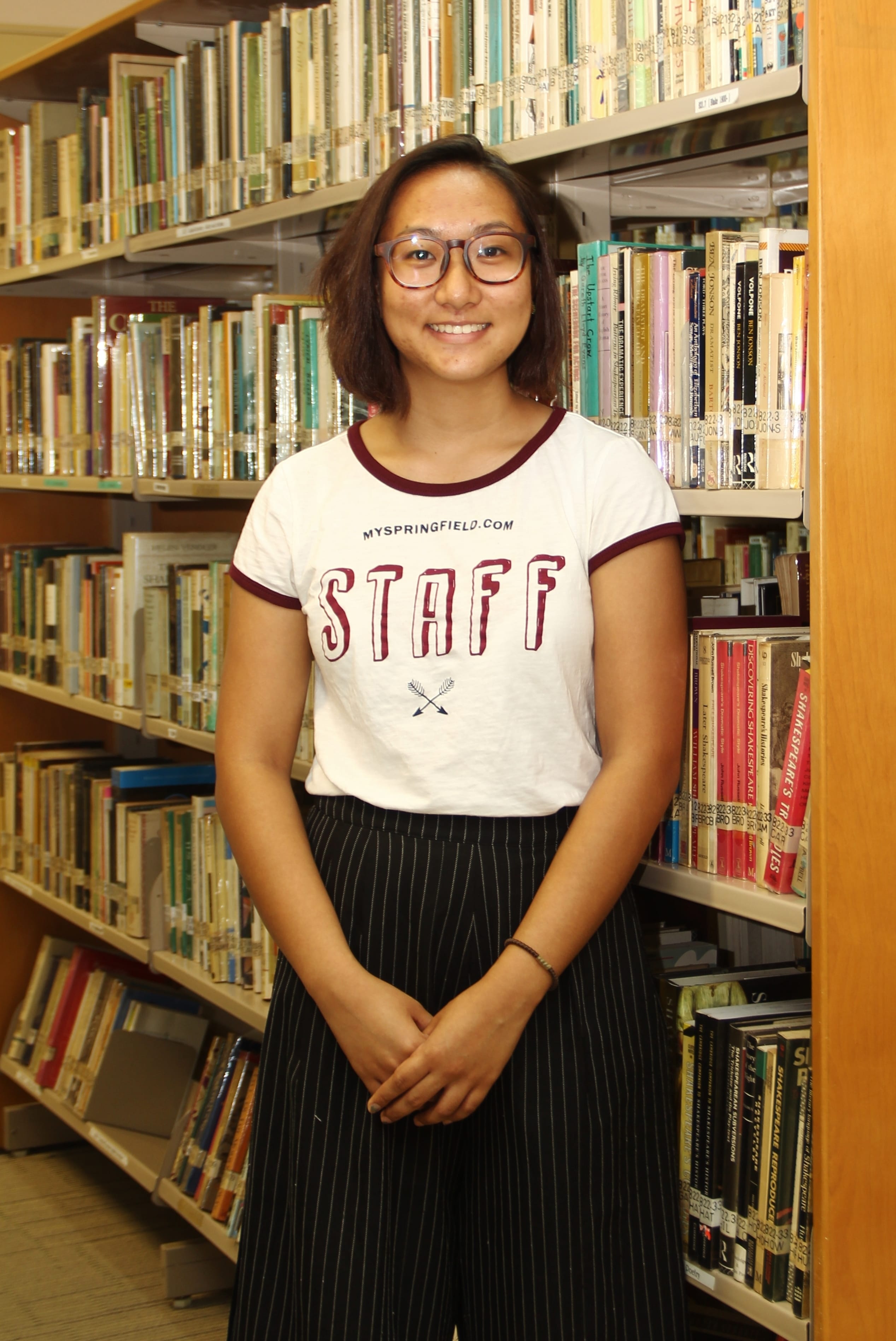 Young woman with glasses wearing a white staff t-shirt stands smiling between library bookshelves.
