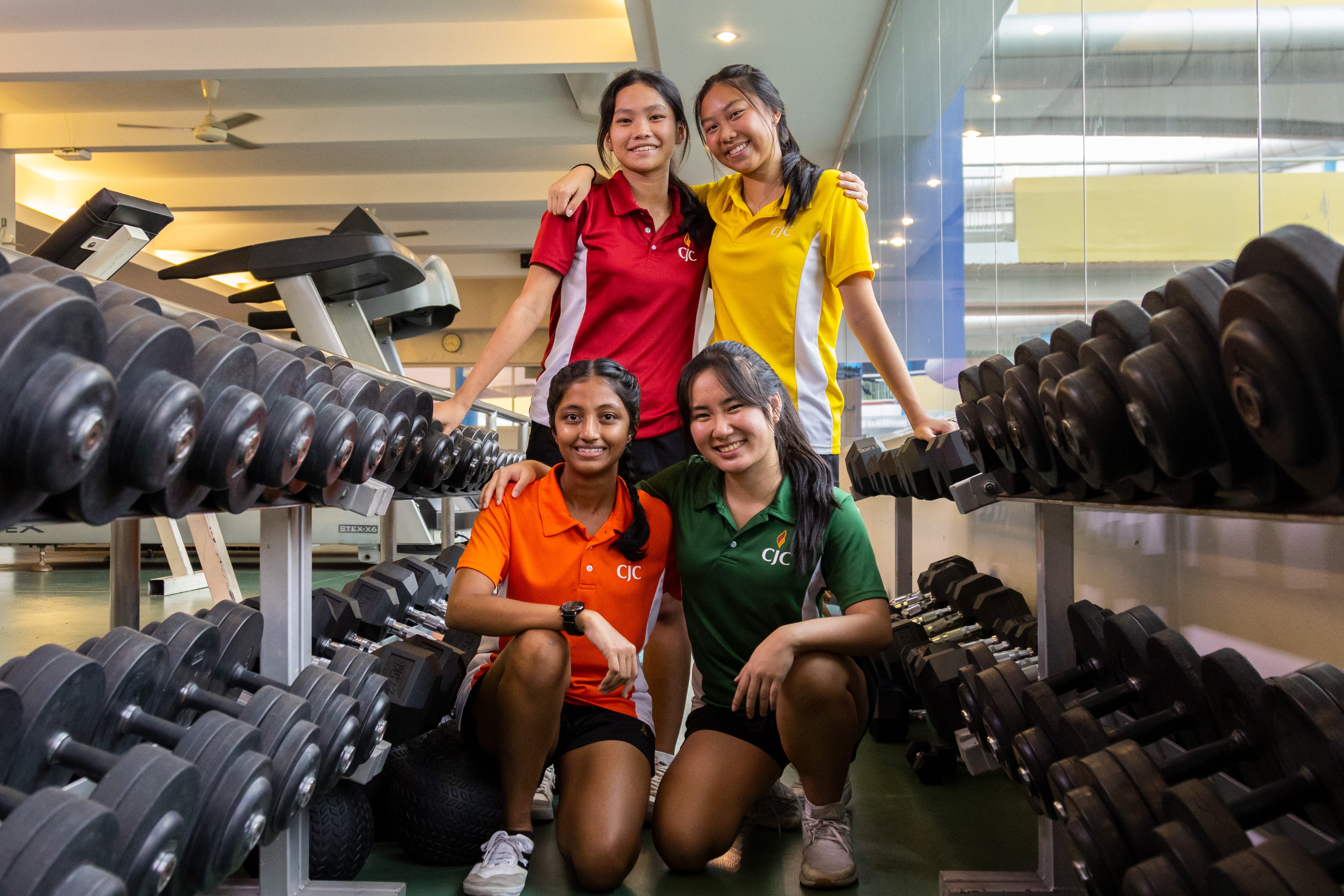 Four girls in colored polo shirts pose smiling between racks of dumbbells in a gym.