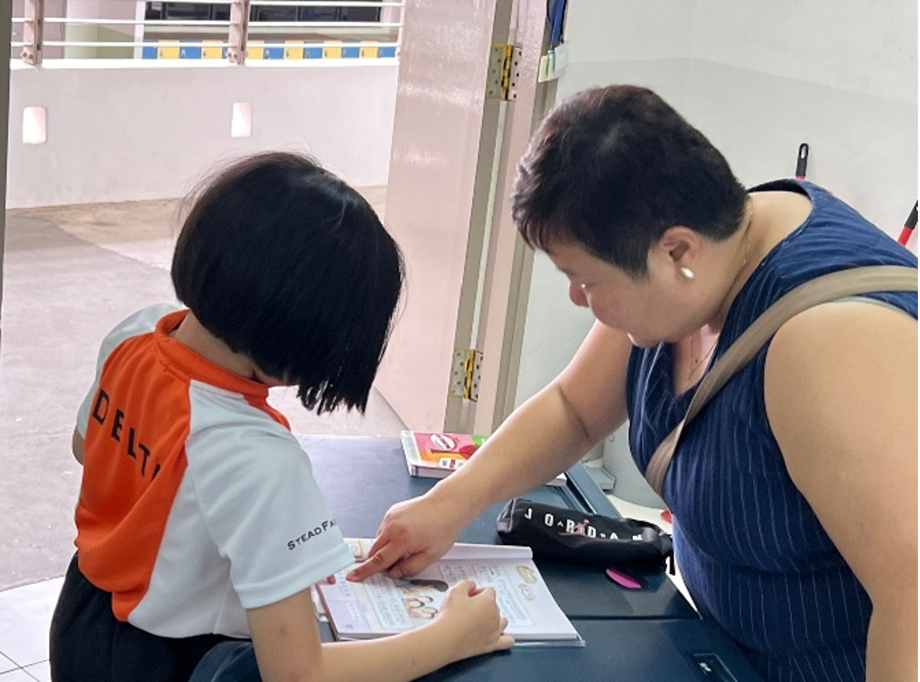 An adult points at a page in a book while a child in an orange and white shirt looks on at a desk.