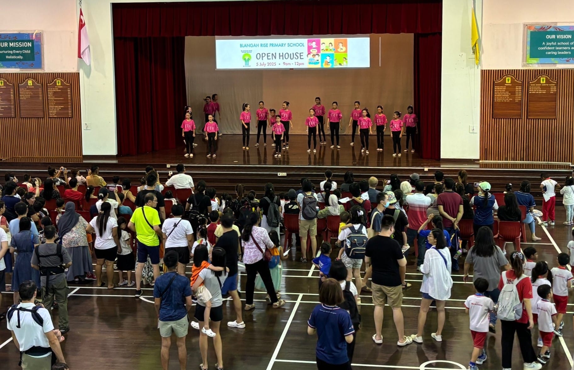 Crowd watches children in pink shirts performing on a school stage beneath an Open House banner.