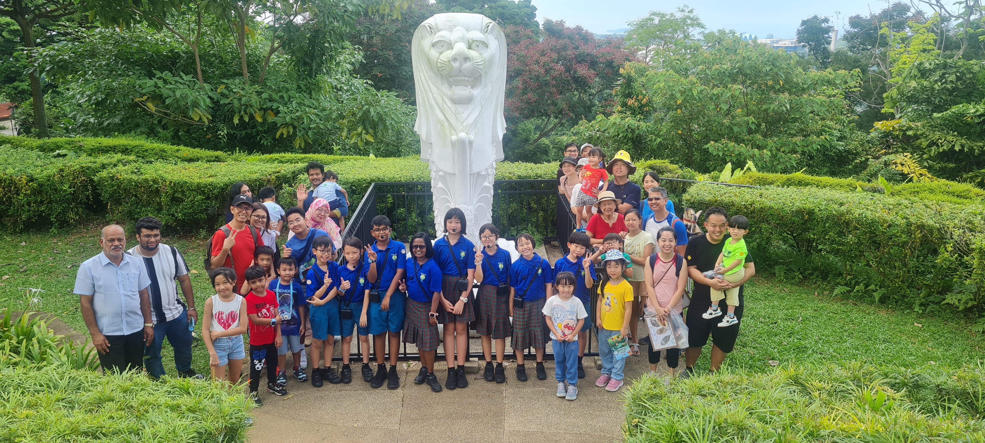 A large group of adults and children in blue uniforms pose in front of a white Merlion statue surrounded by greenery.