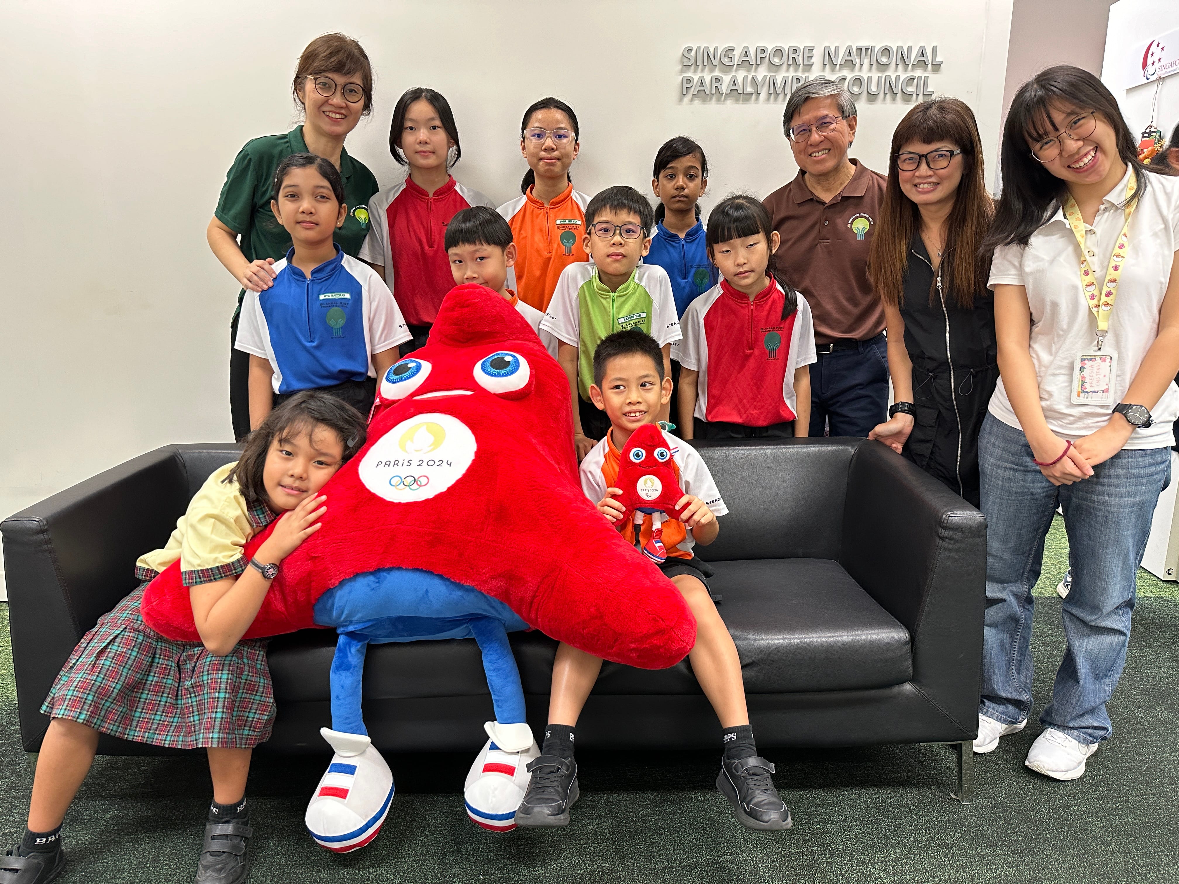 Group of adults and children posing indoors with a large red Paris 2024 plush on a black sofa.