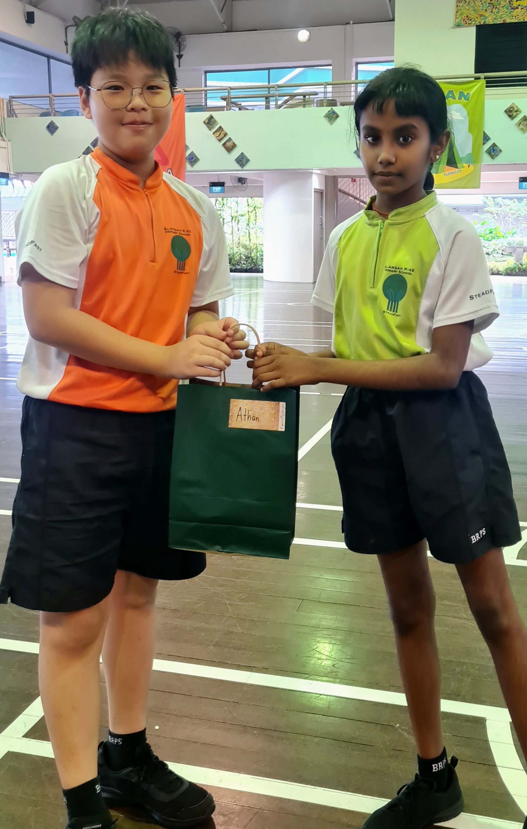 Two children in school uniforms hold a green gift bag labeled "Athan" together in a school hall.