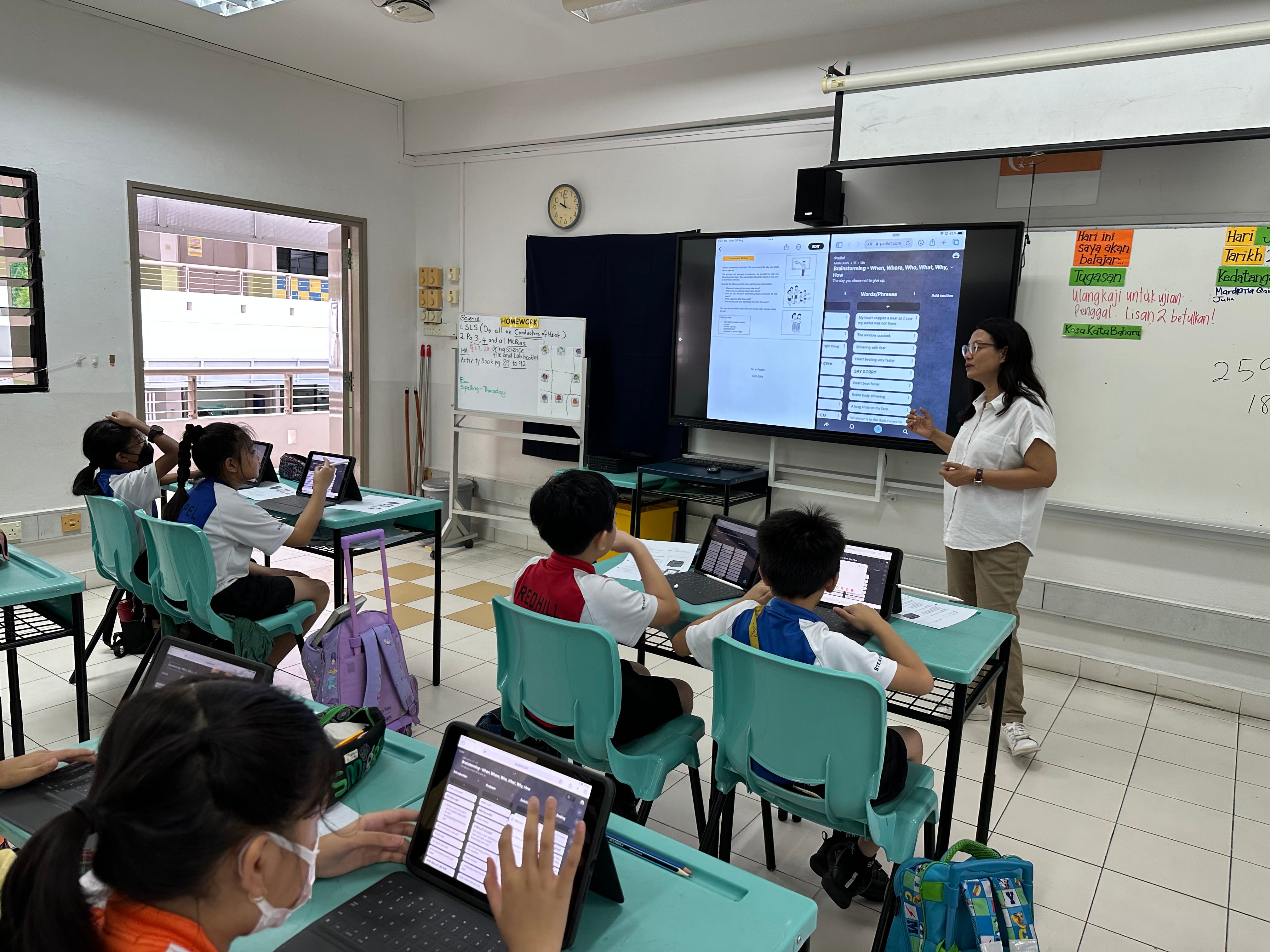 A teacher stands by a large screen while young students sit at desks using tablets in a bright classroom.