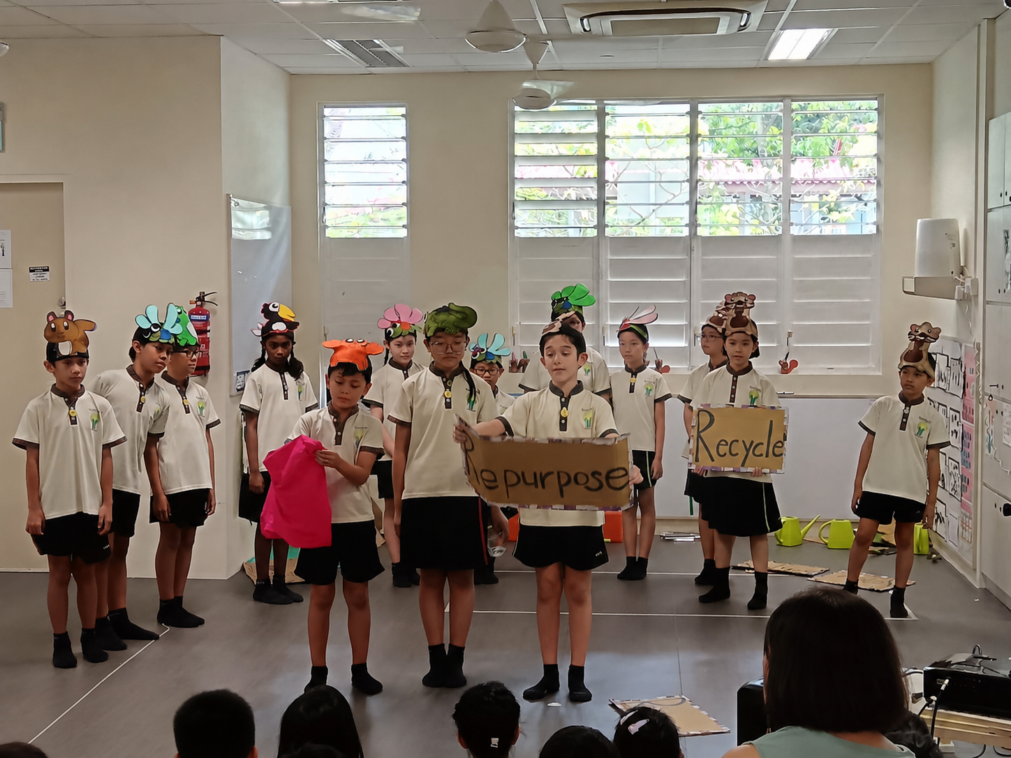 Schoolchildren in animal hats perform, holding signs reading Repurpose and Recycle in a classroom.