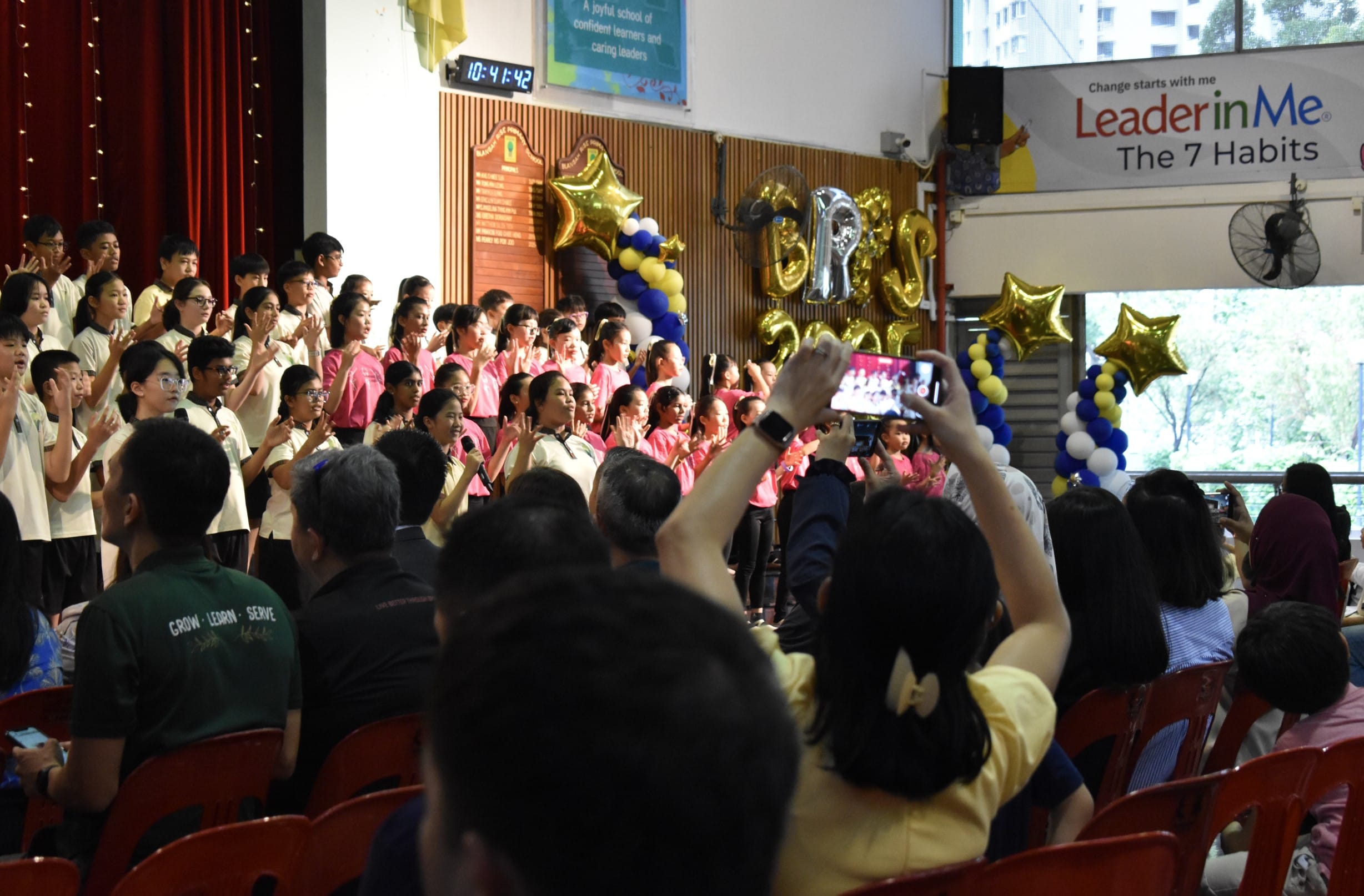 Students in white and pink shirts perform on stage at a school event with gold balloons, while audience members record with phones.