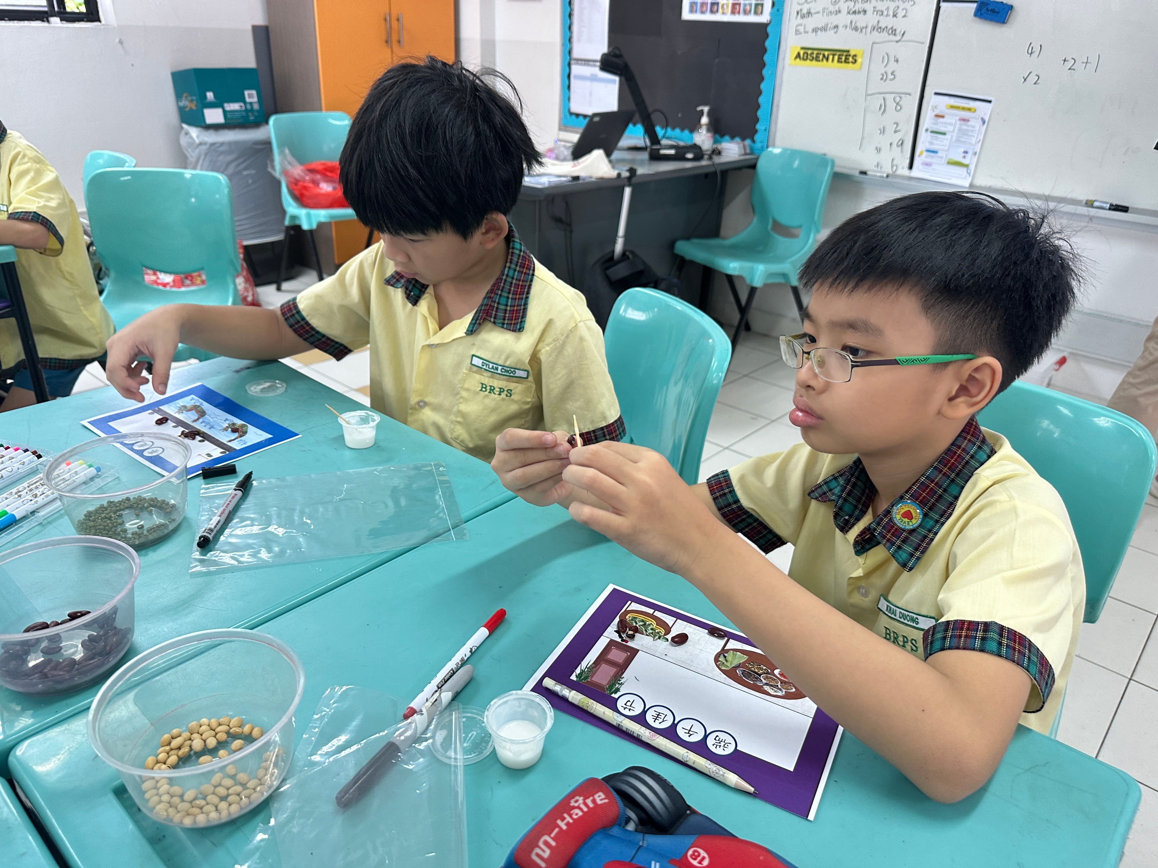 Two young students in yellow school uniforms work on a hands-on activity with seeds at a teal classroom table.
