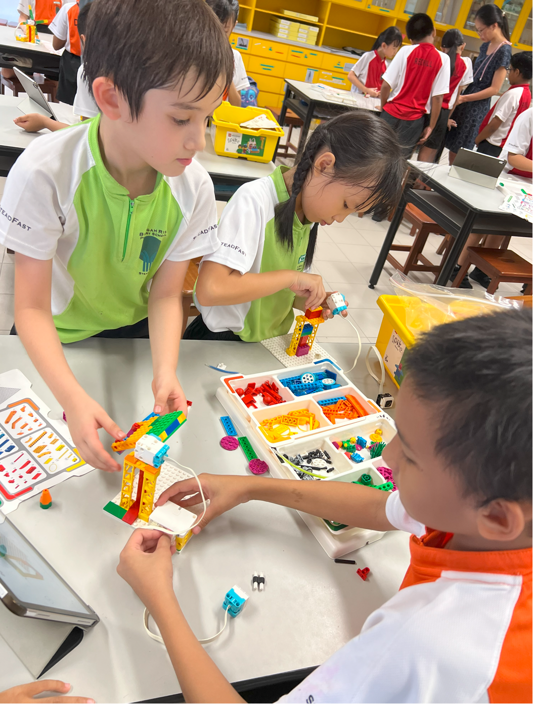 Three school children in uniforms build colorful LEGO models together at a classroom table.