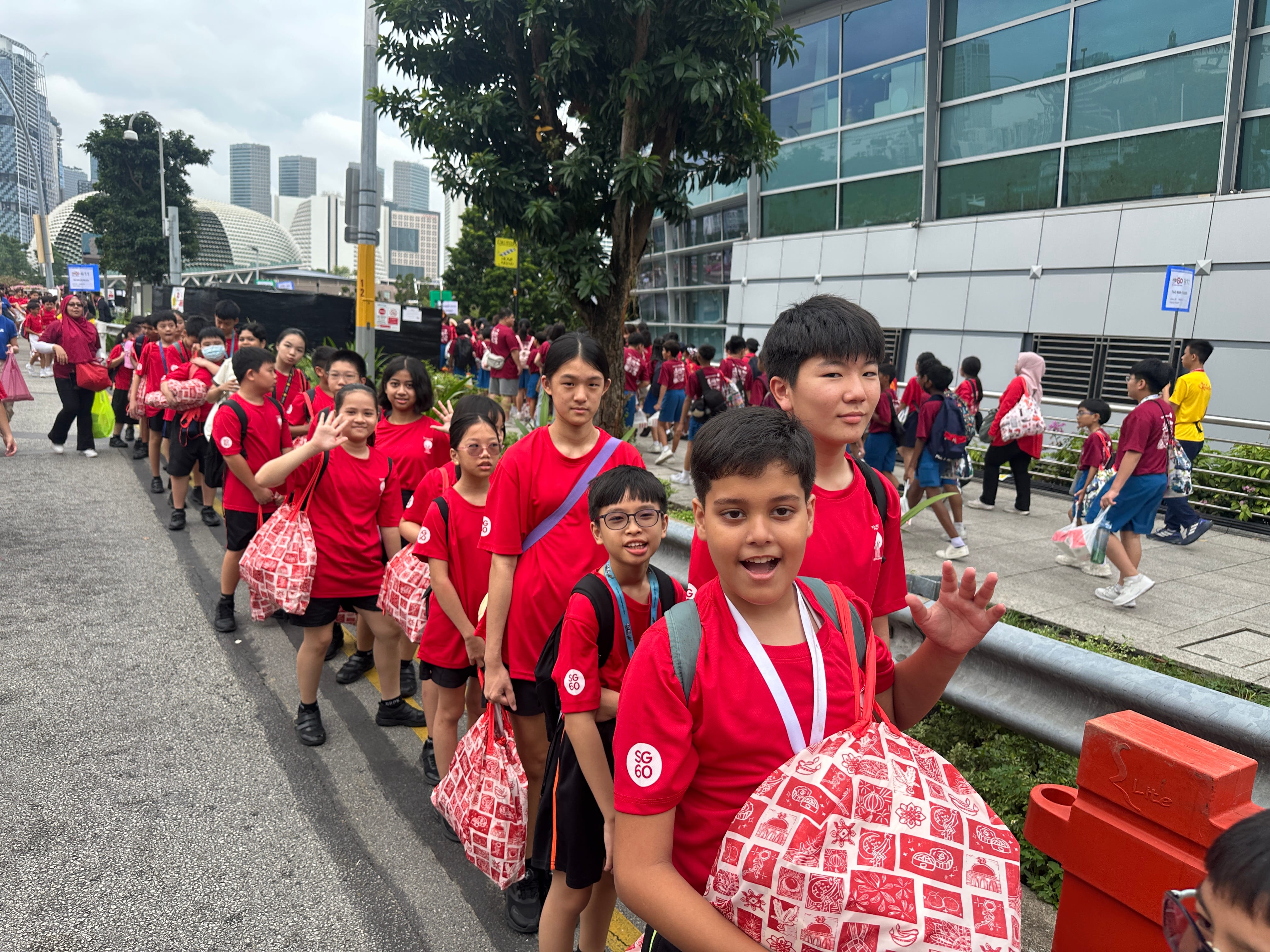 A long line of children in red SG60 shirts walk along a city street, some waving and carrying patterned bags.
