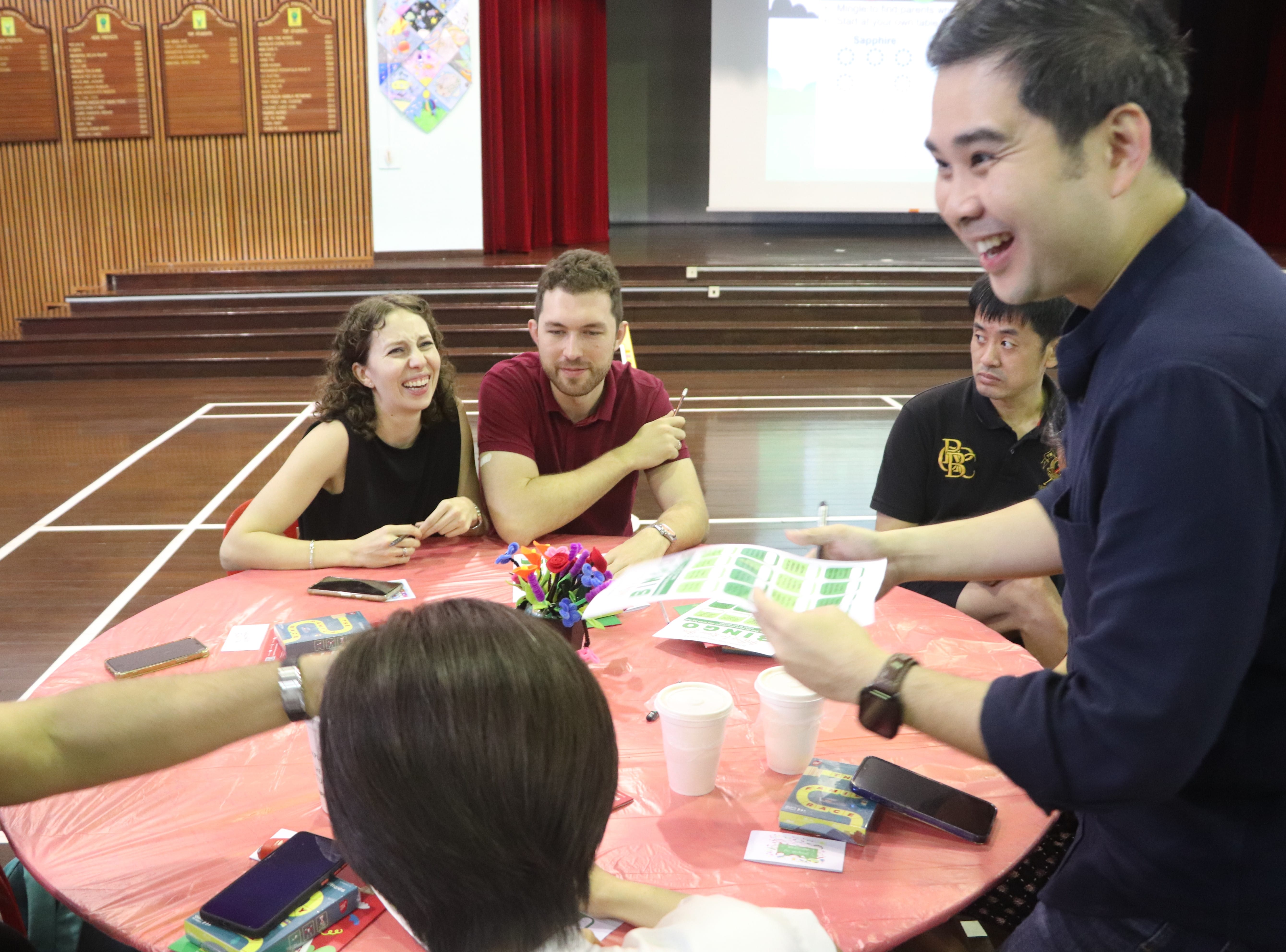 Four people sit around a red table laughing, with papers, phones, and cups on it, in a hall with a projector screen.