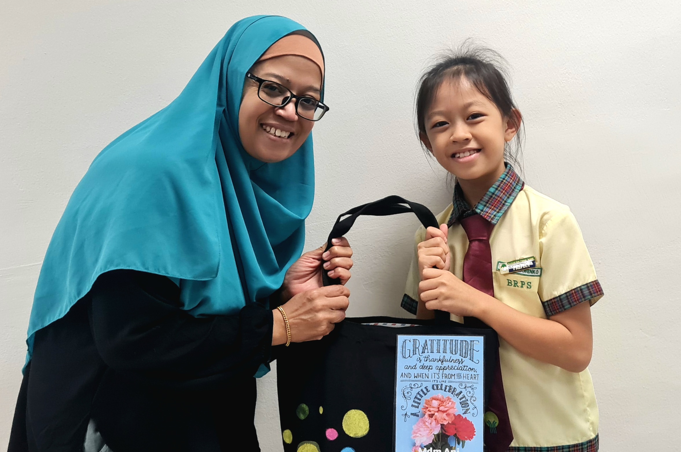 A smiling woman in a teal hijab and a young girl in a school uniform hold a black tote bag with a gratitude quote.