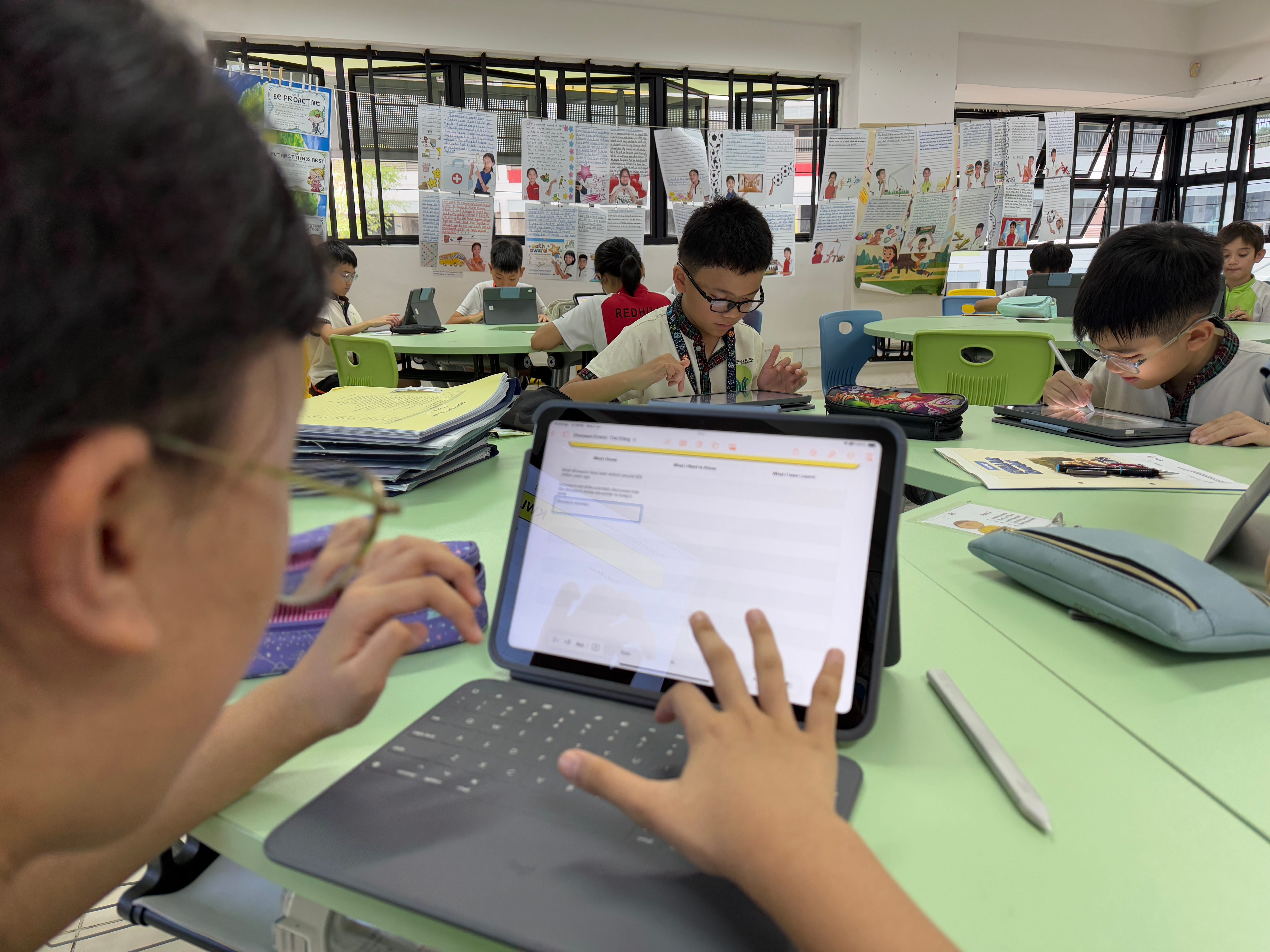 Students in a classroom work on tablets at green tables, with posters covering the back wall.