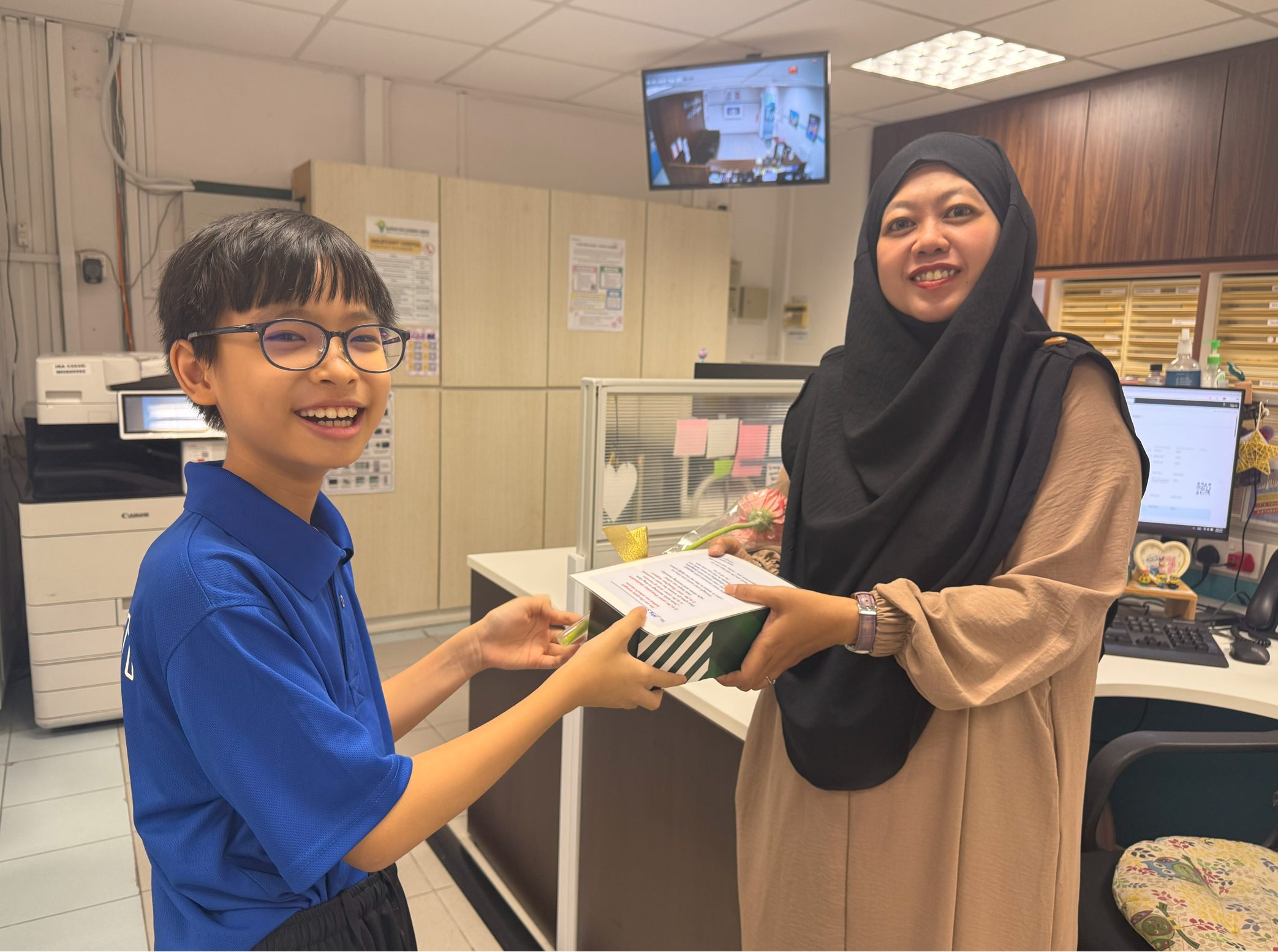 Smiling boy in blue shirt hands a striped box to a smiling woman in hijab at an office counter.