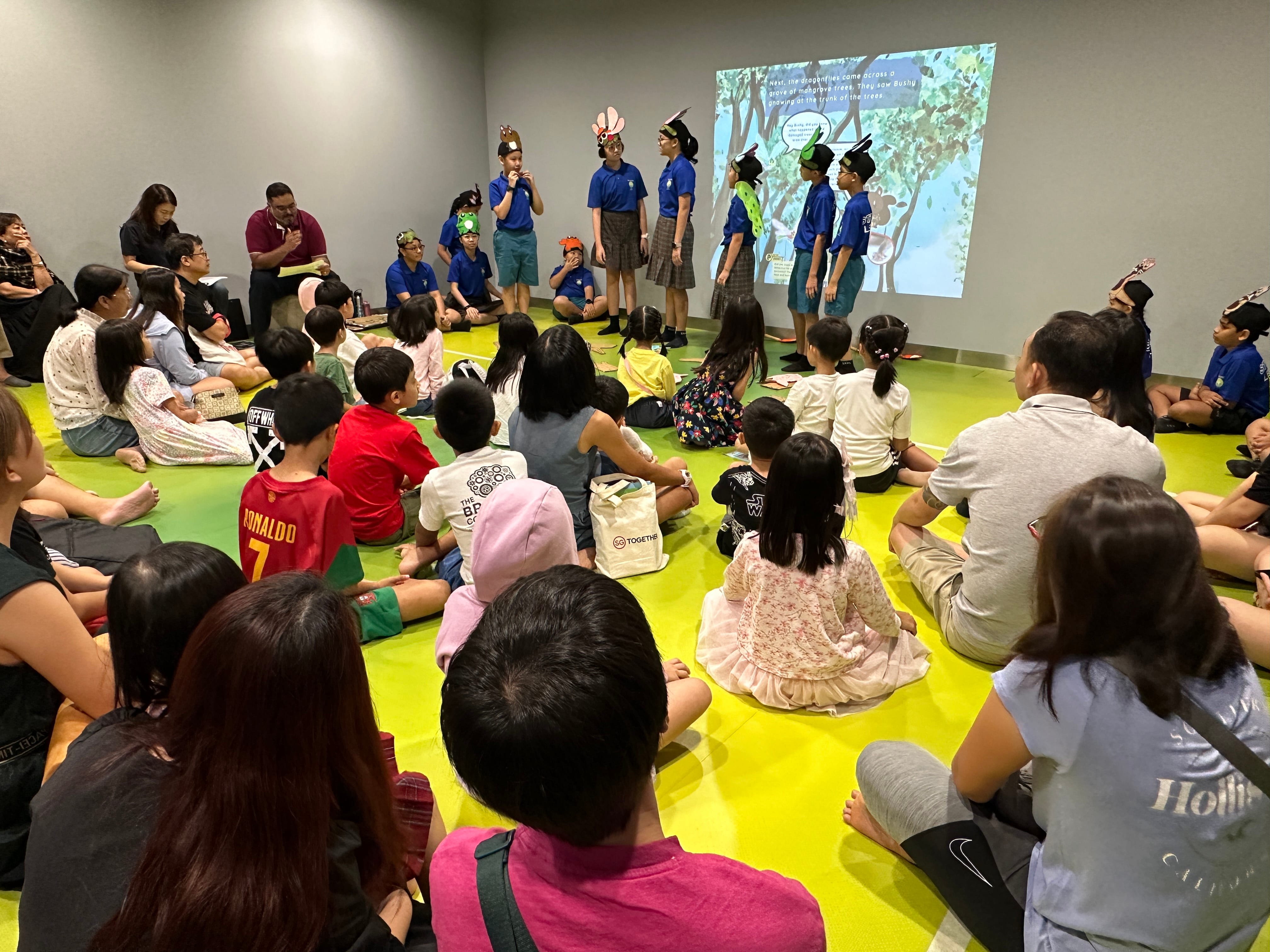 Children seated on floor watch kids in blue shirts wearing animal ears perform before a projected slide.