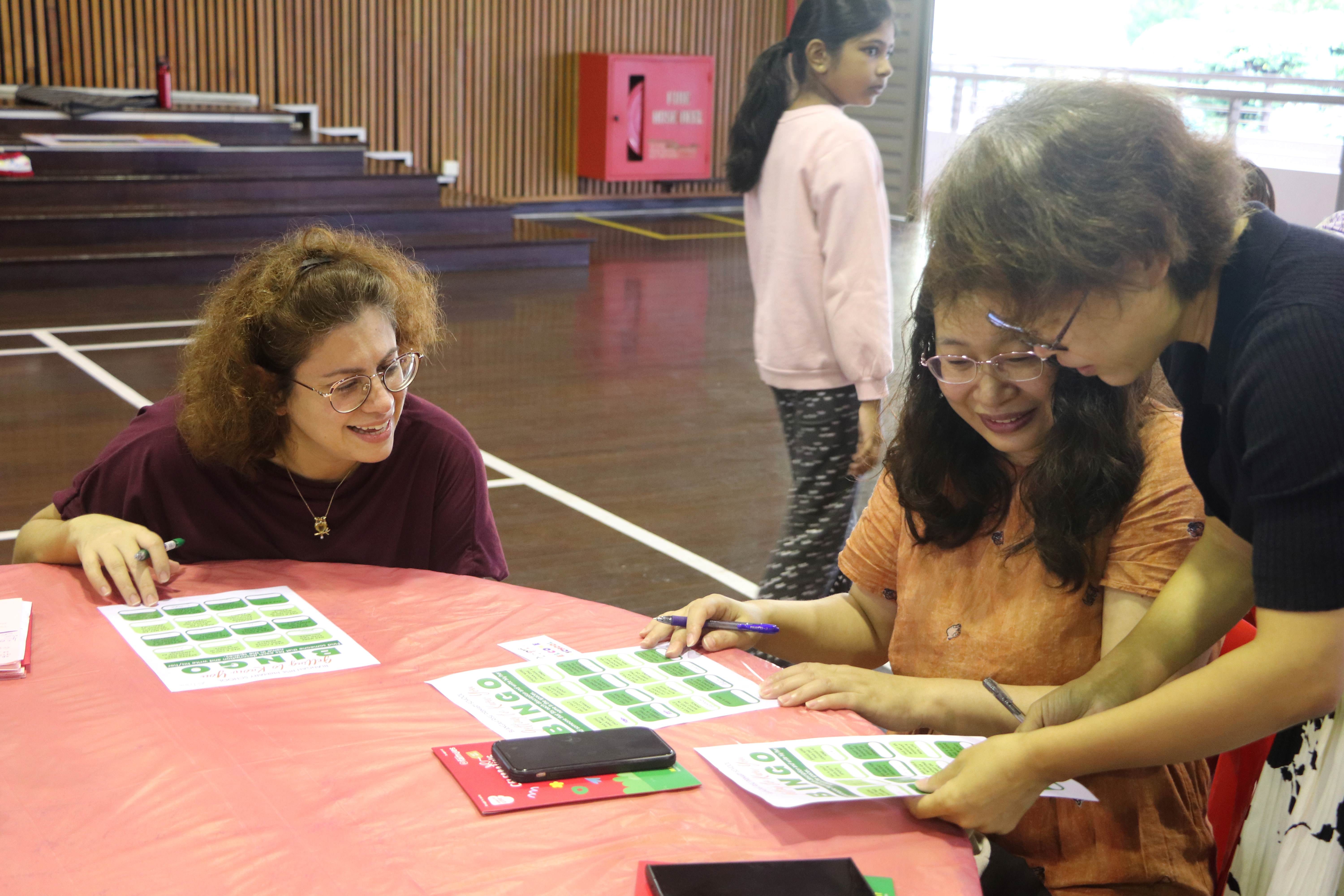 Three people smile around a pink table covered with green and white bingo cards in a hall.