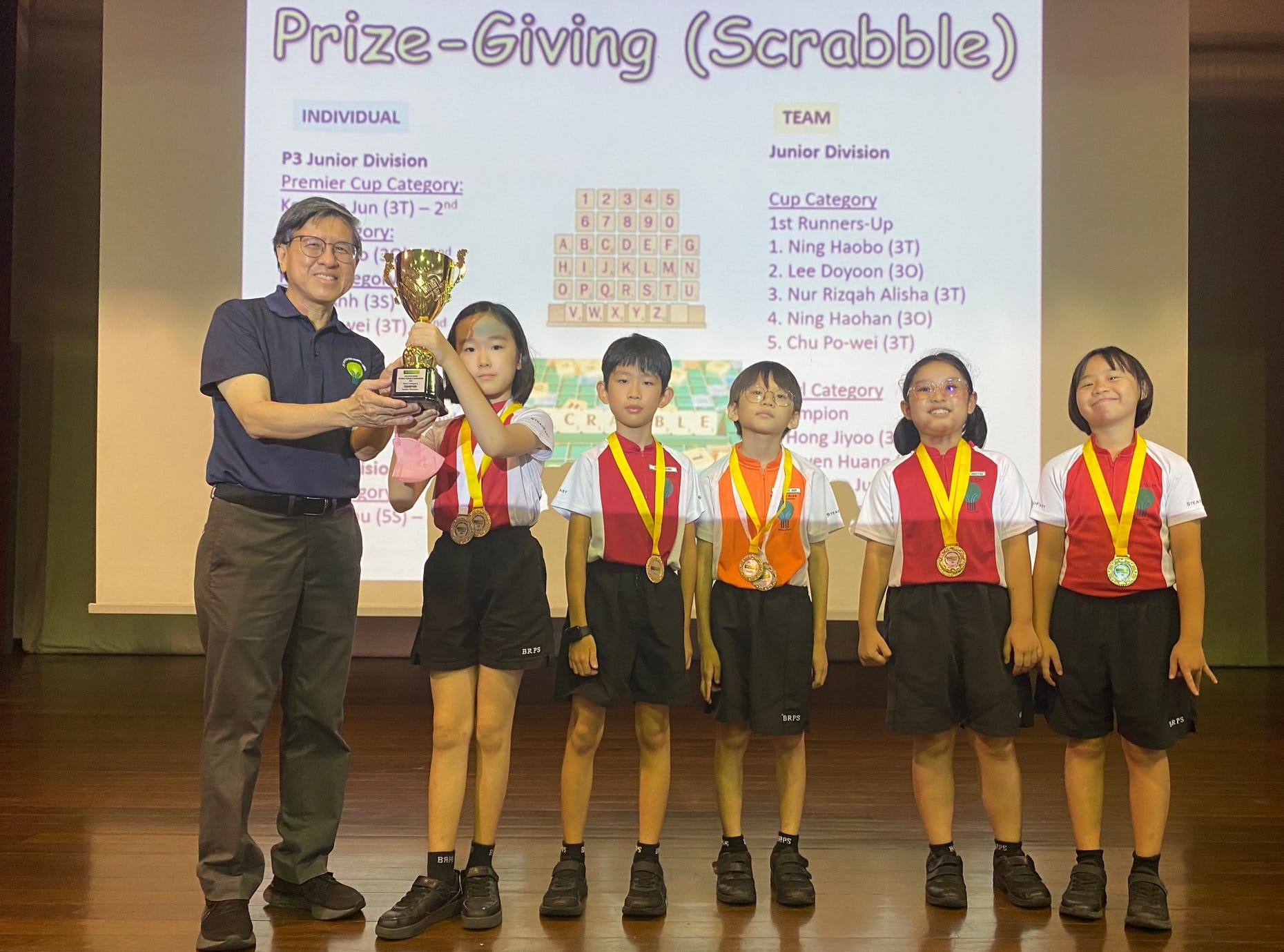 An adult presents a trophy to five medal-wearing children at a Scrabble Prize-Giving ceremony on a stage.