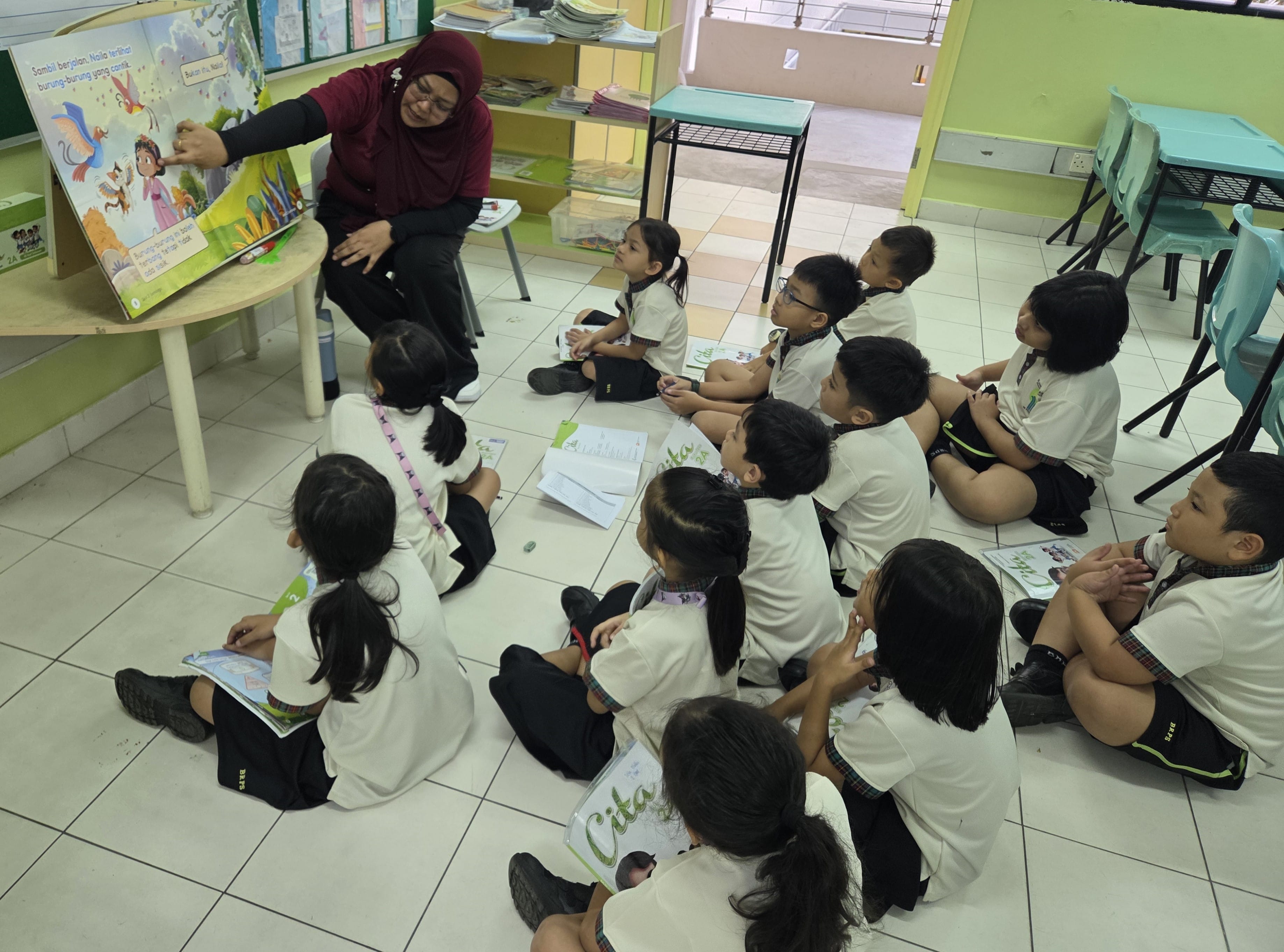 Teacher in hijab reads a picture book to seated children in uniform on a classroom floor.