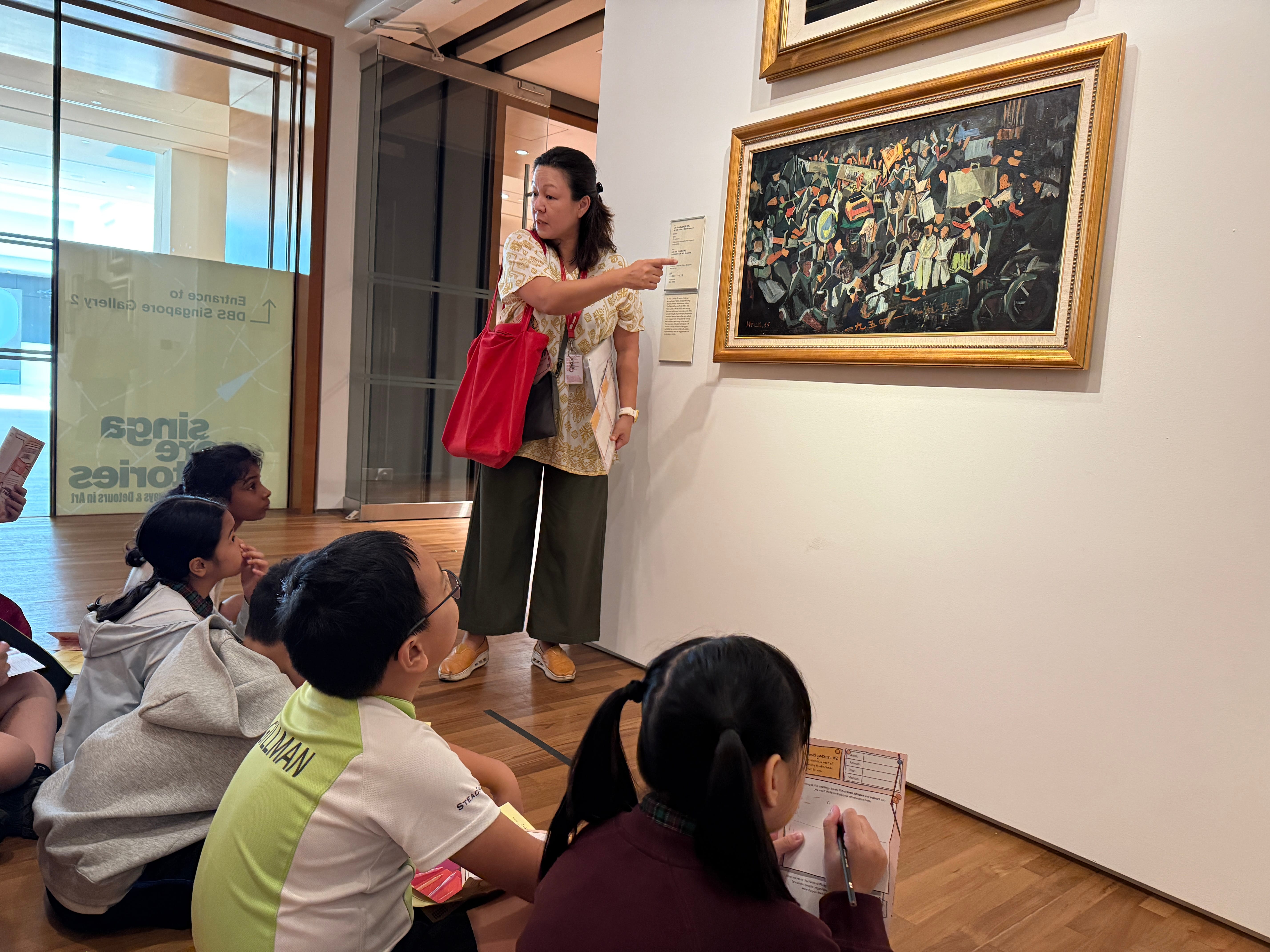Museum guide points at framed painting while seated children watch and take notes.