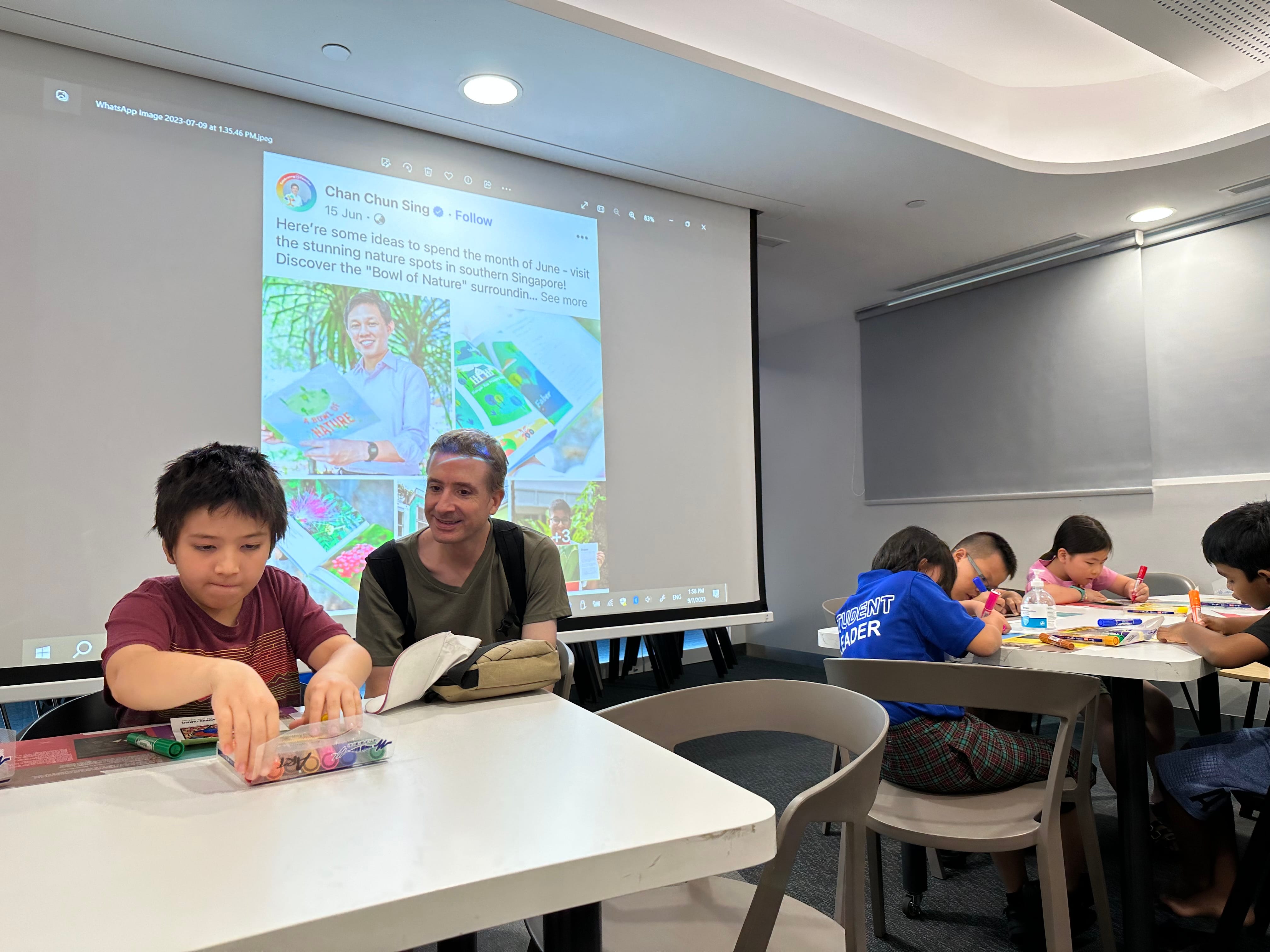 Children and an adult sit at white tables drawing, with a projected social media post visible on a screen behind them.