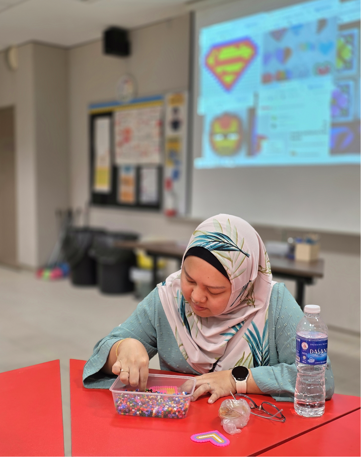 Woman wearing a hijab sorts colorful beads in a container at a red table in a classroom with a projector screen.