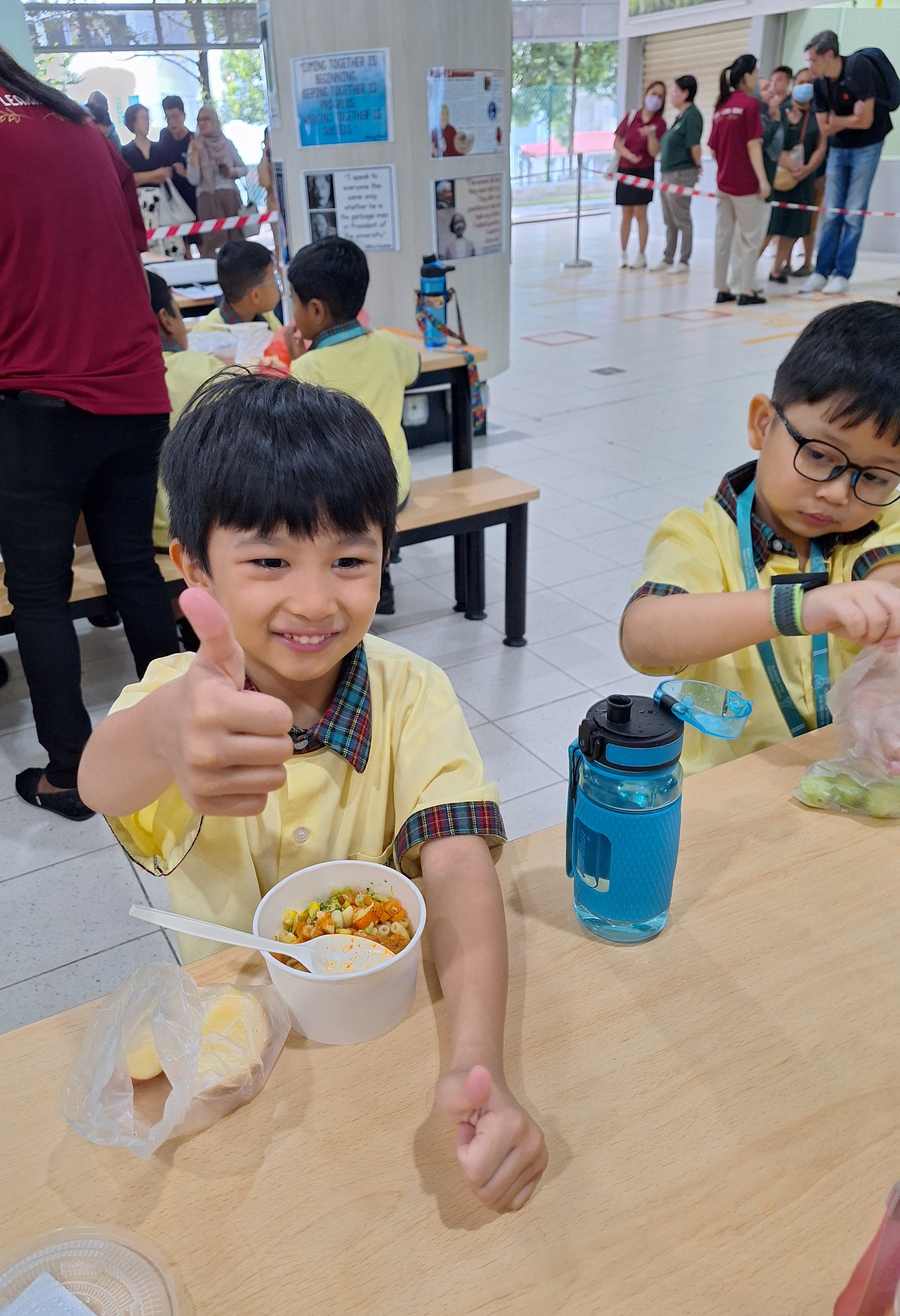 A smiling young boy in a yellow school uniform gives two thumbs up at a cafeteria table with a bowl of food.