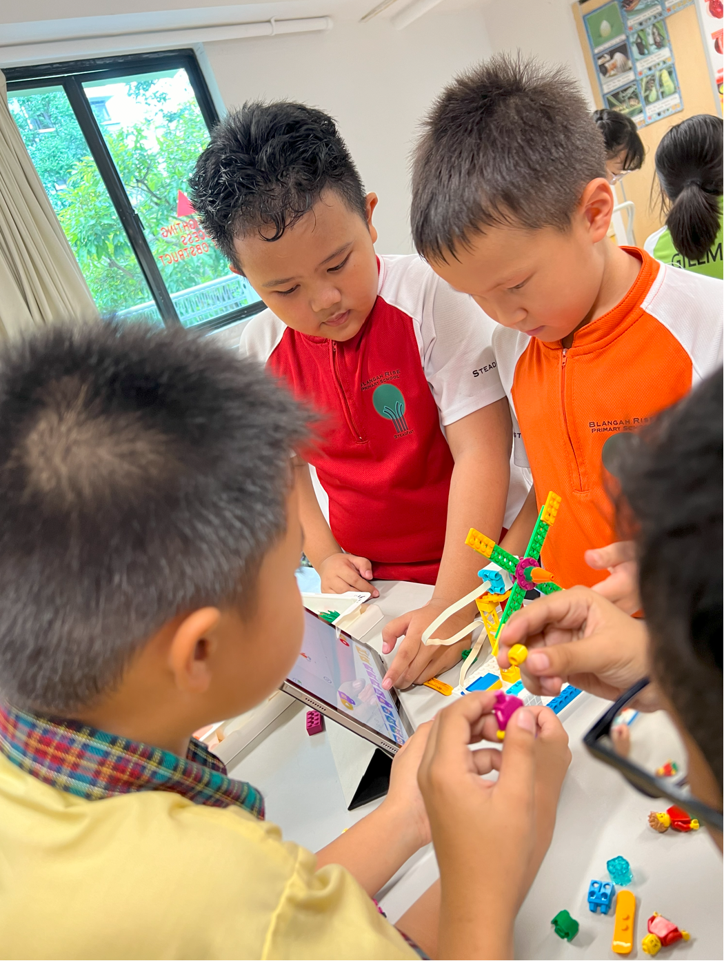 Four young students in school uniforms gather around a table, building a colorful plastic brick structure using a tablet as reference.