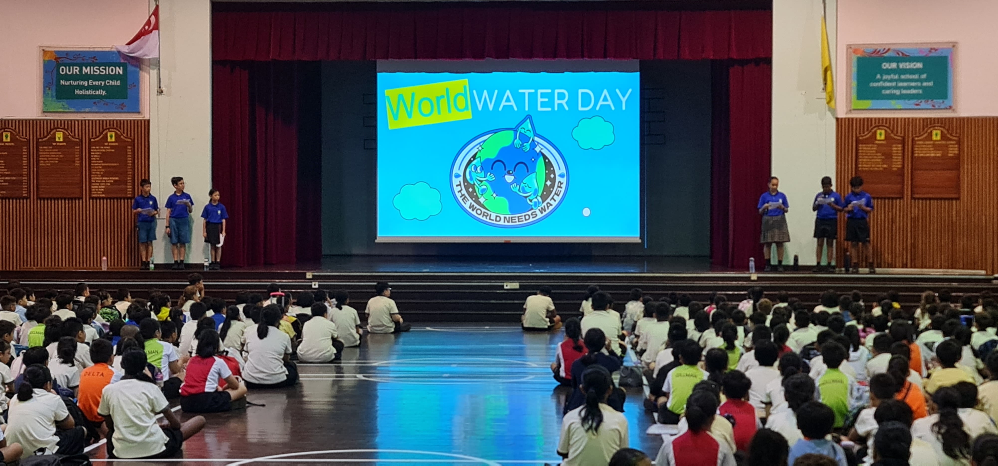 Students sit on a school hall floor facing a stage with a World Water Day presentation on a large screen.