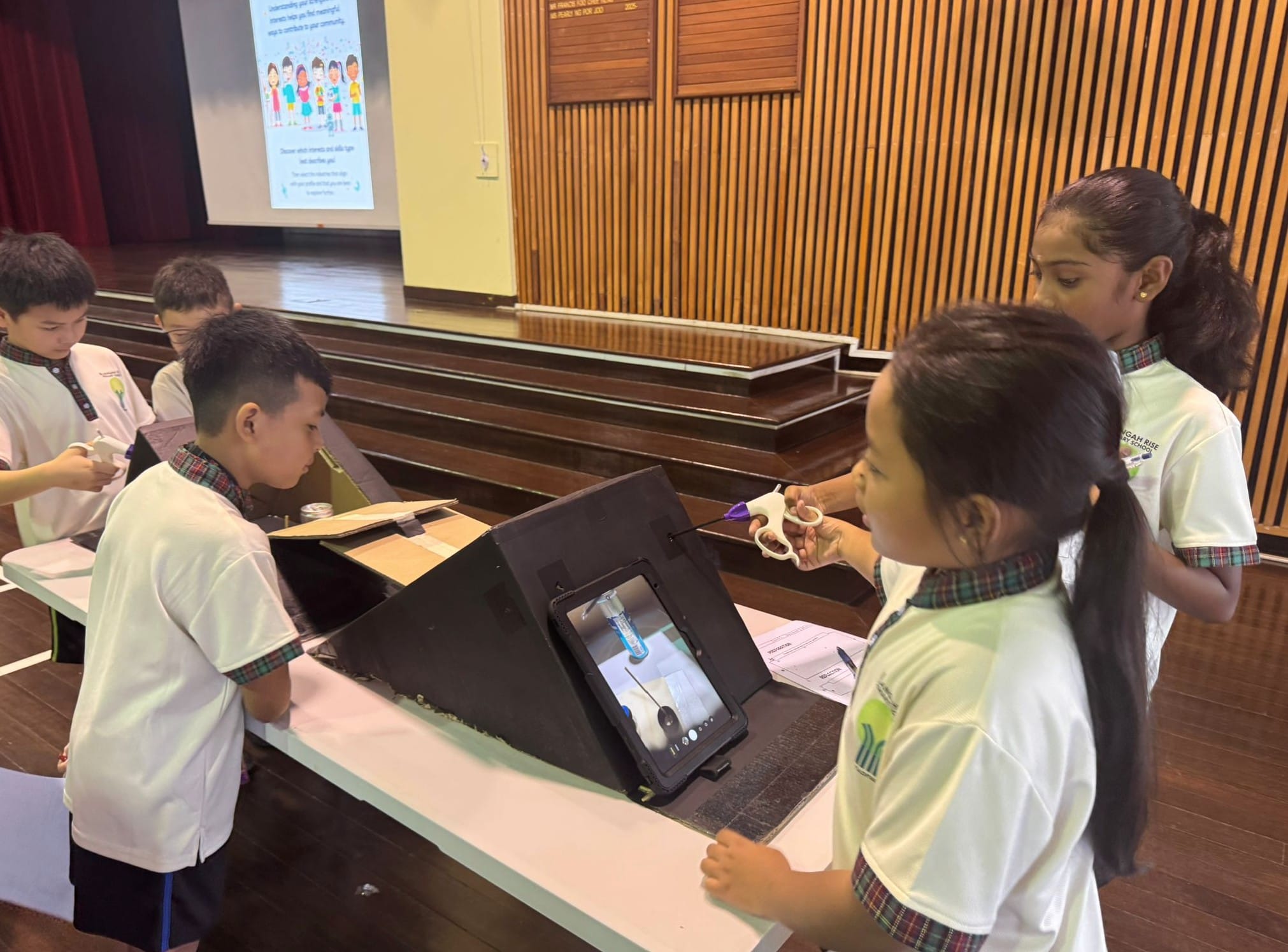Four school children in white uniforms work on a project using a tablet and small tools at a table in a hall.