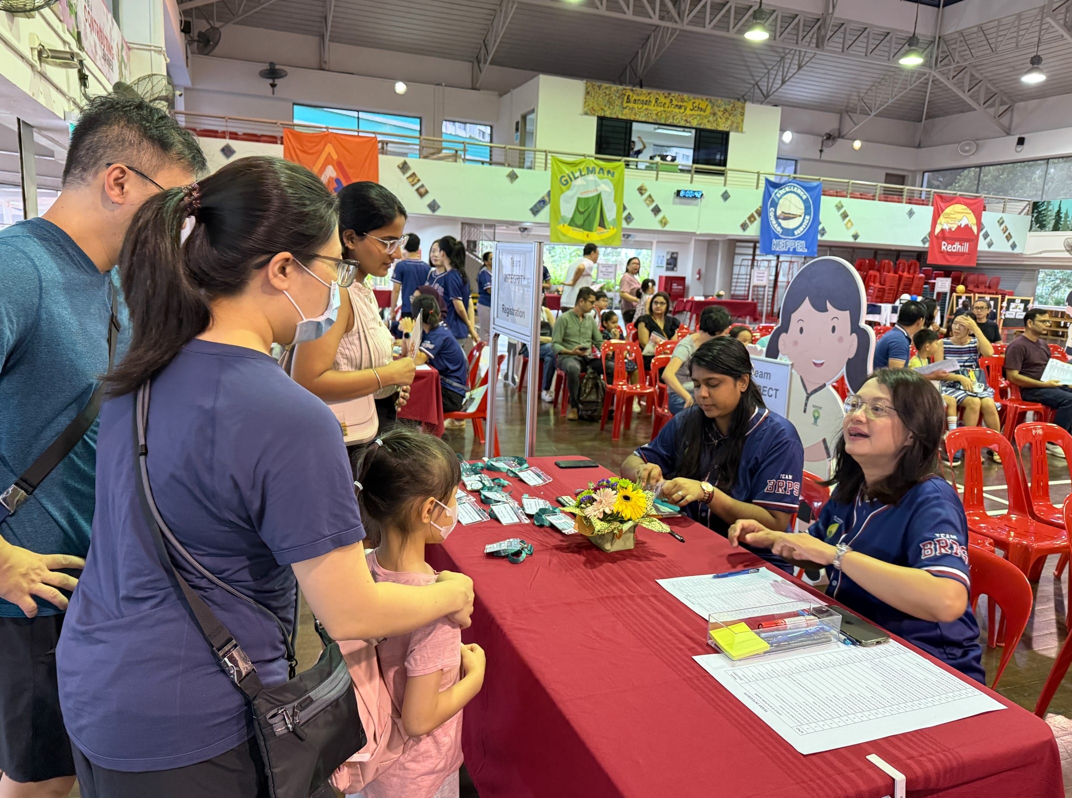 A family registers at a red-clothed table staffed by two women in blue jerseys inside a busy school hall.