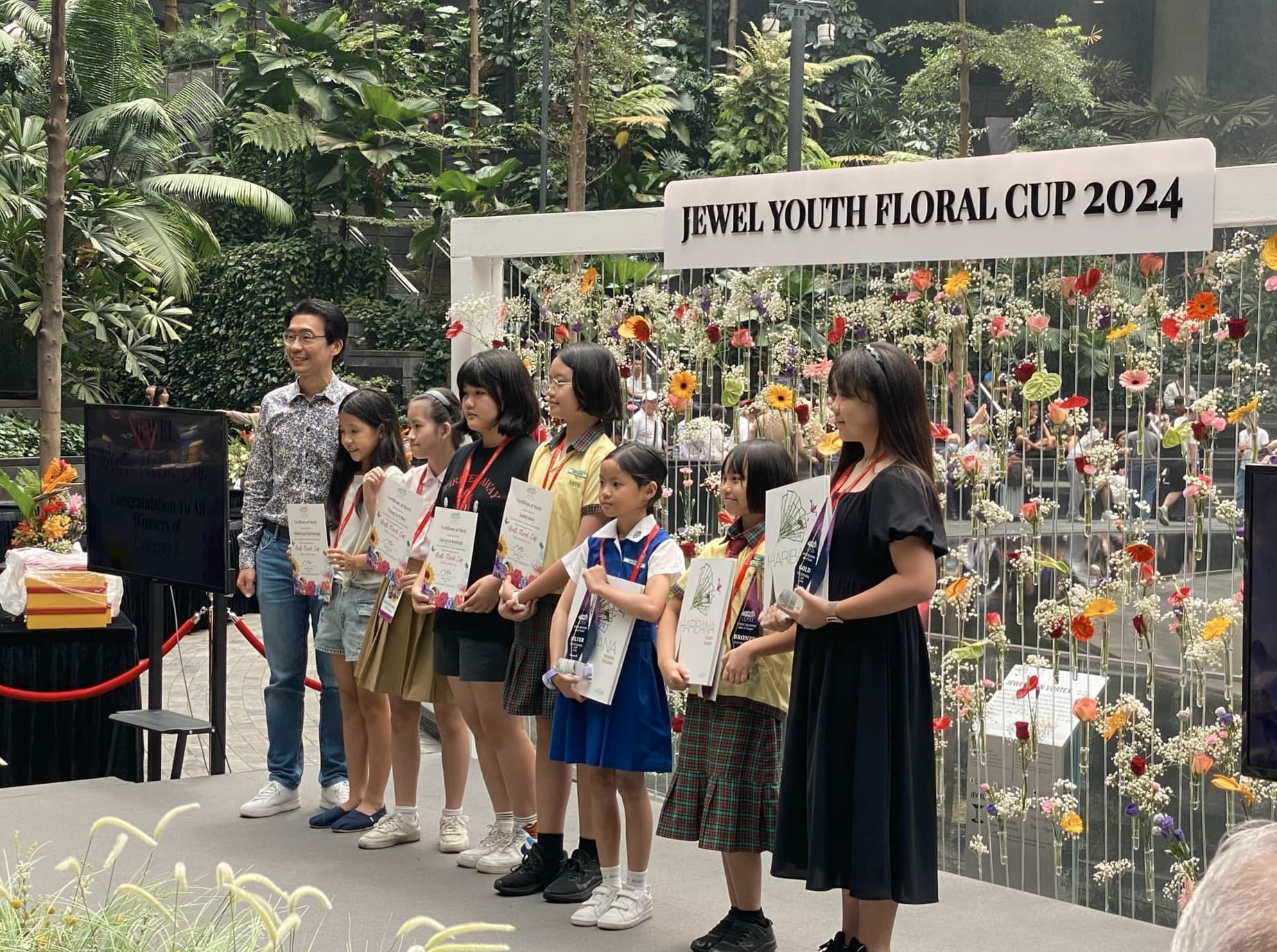 Children and an adult hold certificates on stage at the Jewel Youth Floral Cup 2024, with colorful flowers displayed behind them.