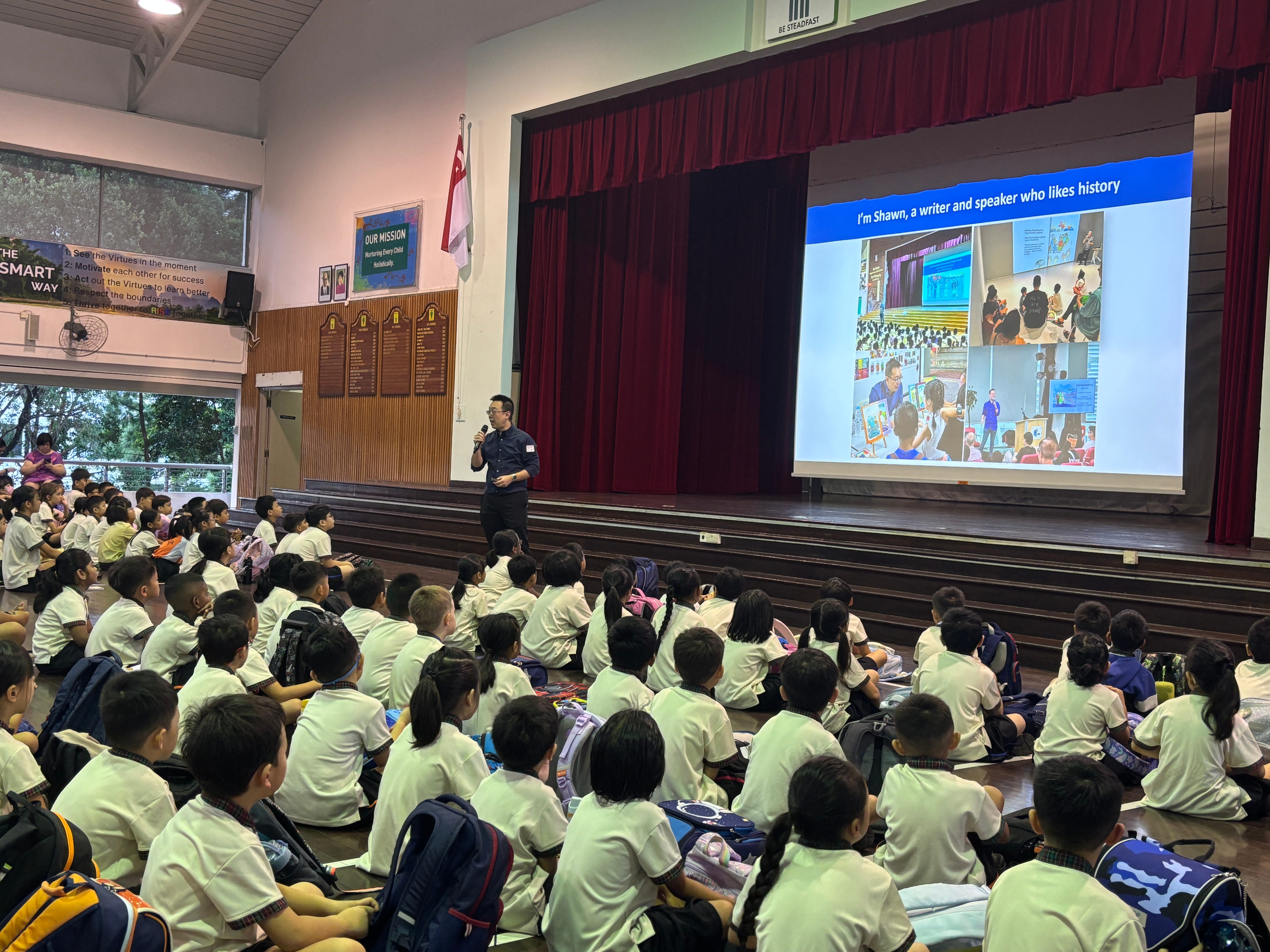 Students sit on the floor in a school hall as a man speaks on stage beside a large projection screen.