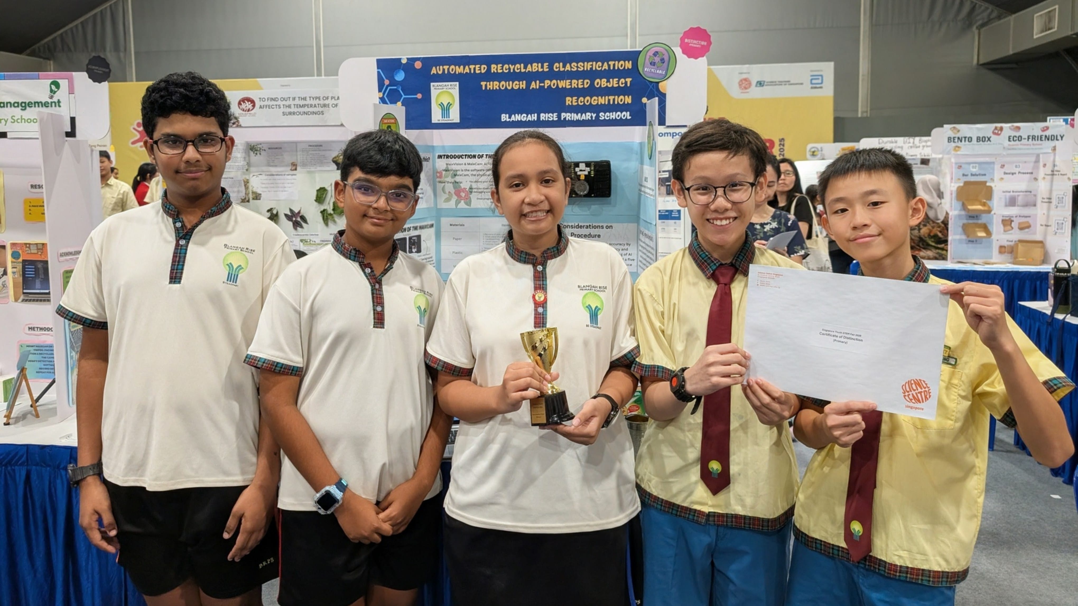 Five primary school students in uniform pose with a trophy and certificate in front of a science fair project board.