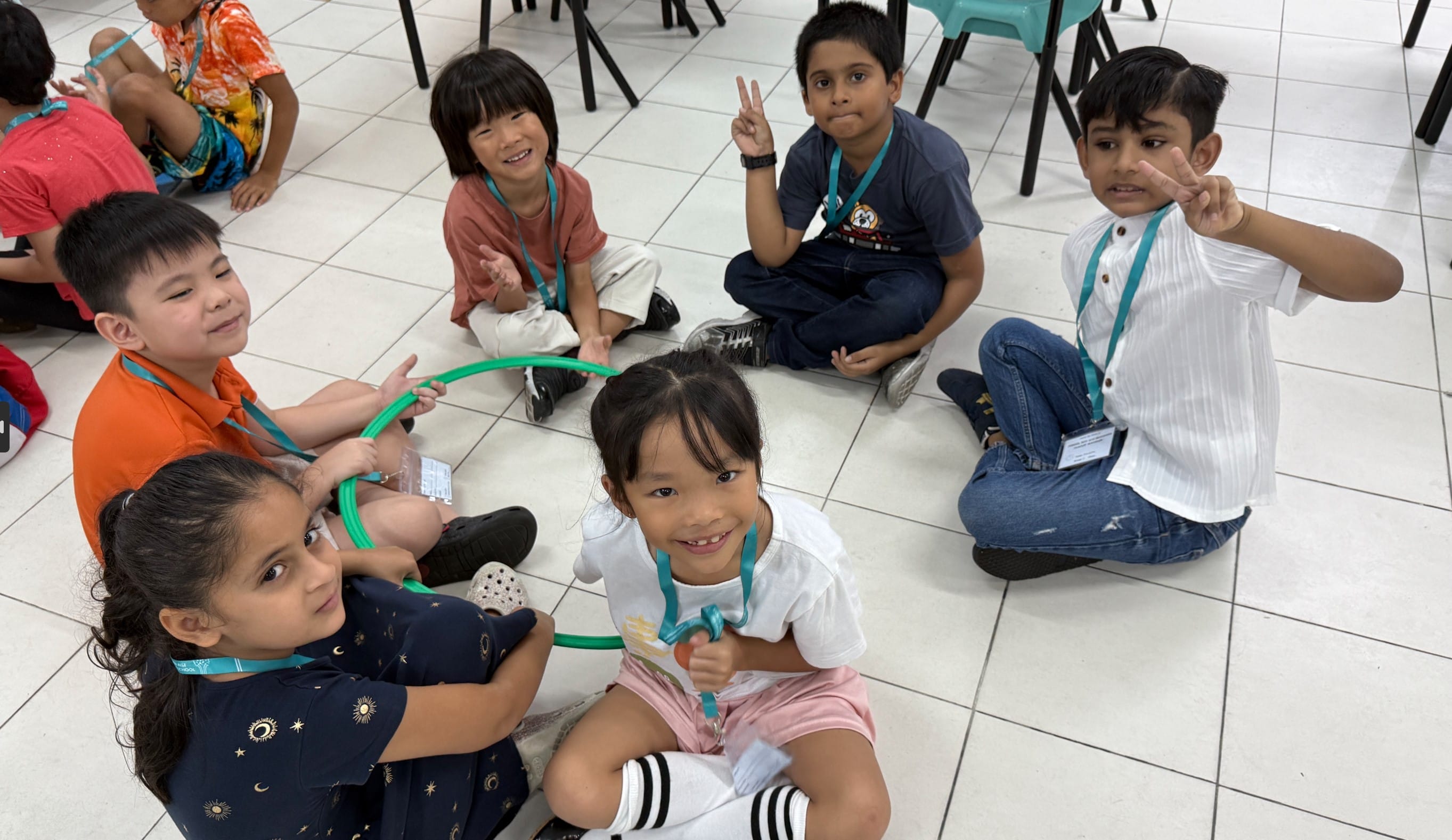 Six young children wearing lanyards sit on a tiled floor smiling, some making peace signs, holding a green hoop.