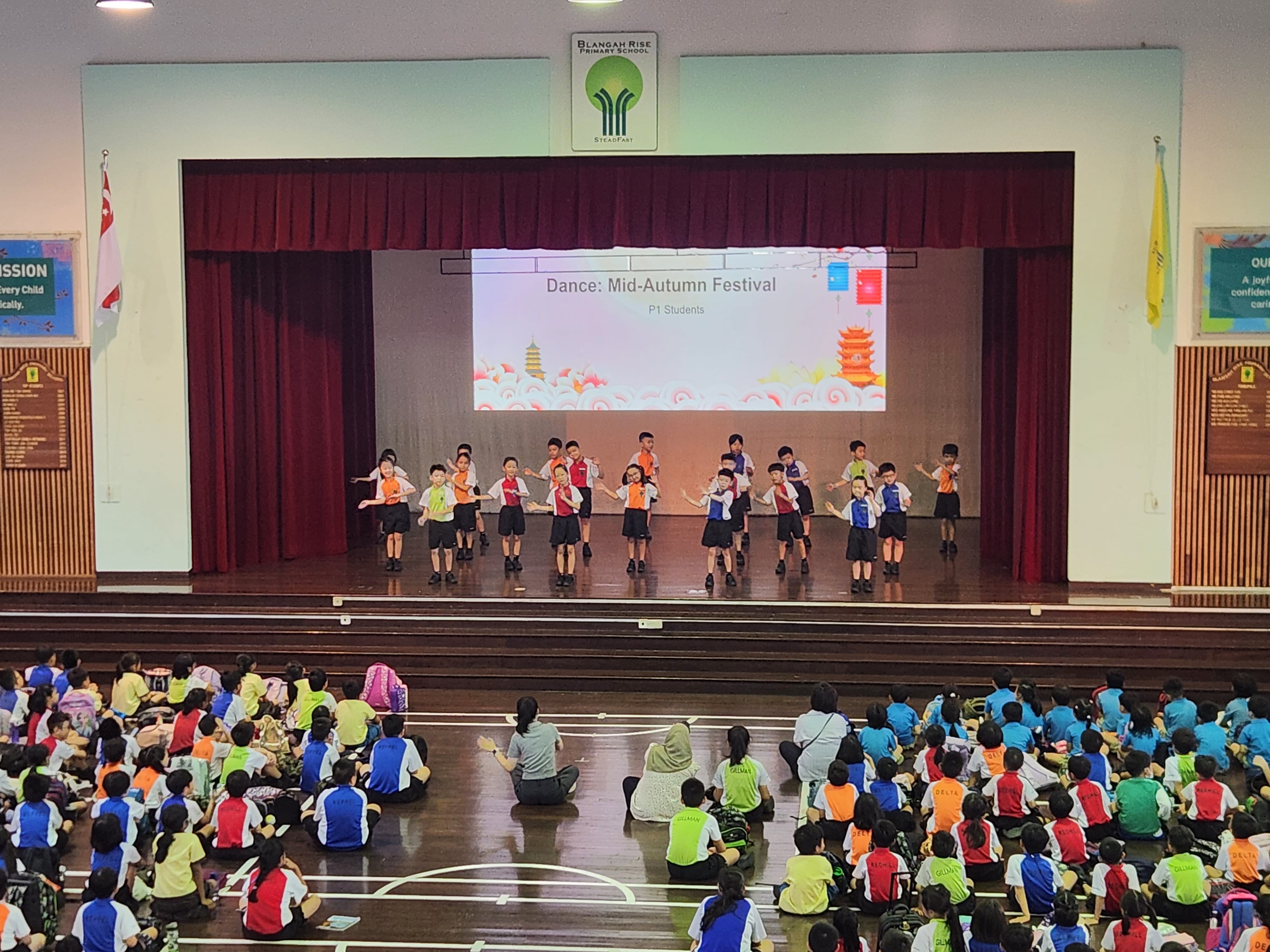 School auditorium with children dancing on stage; seated students watch from the floor.