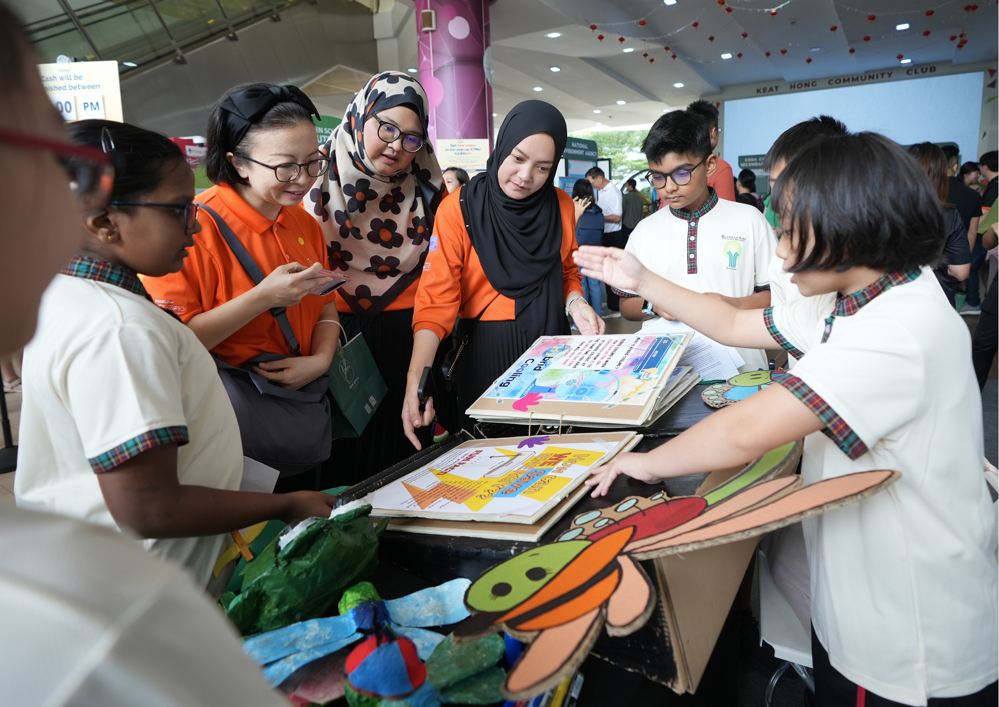 Students and adults gather at a booth, pointing at posters and colorful cardboard insect crafts on a table.