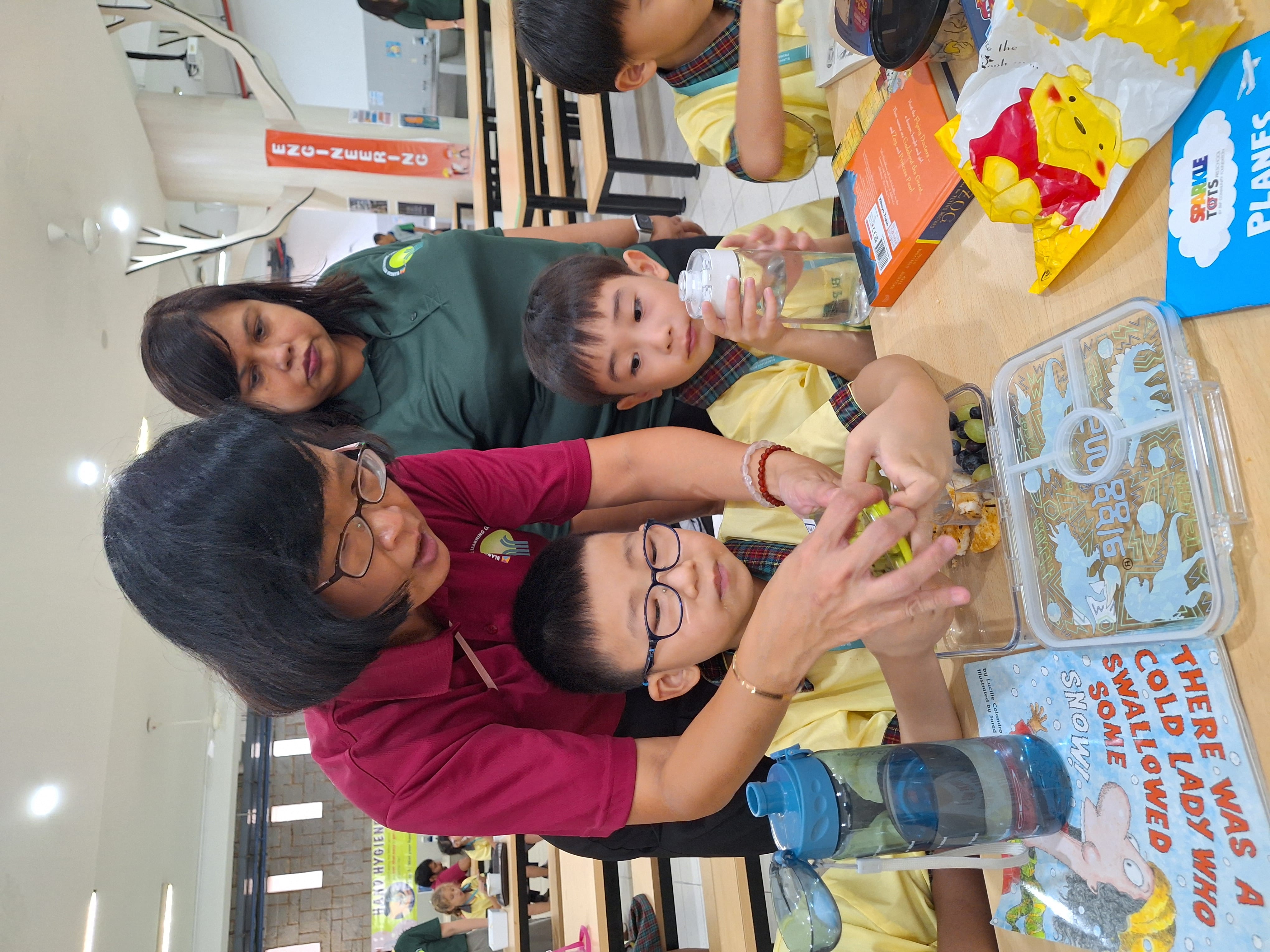Teacher helps two boys open a lunch container at a cafeteria table with books, snacks, and water bottles.