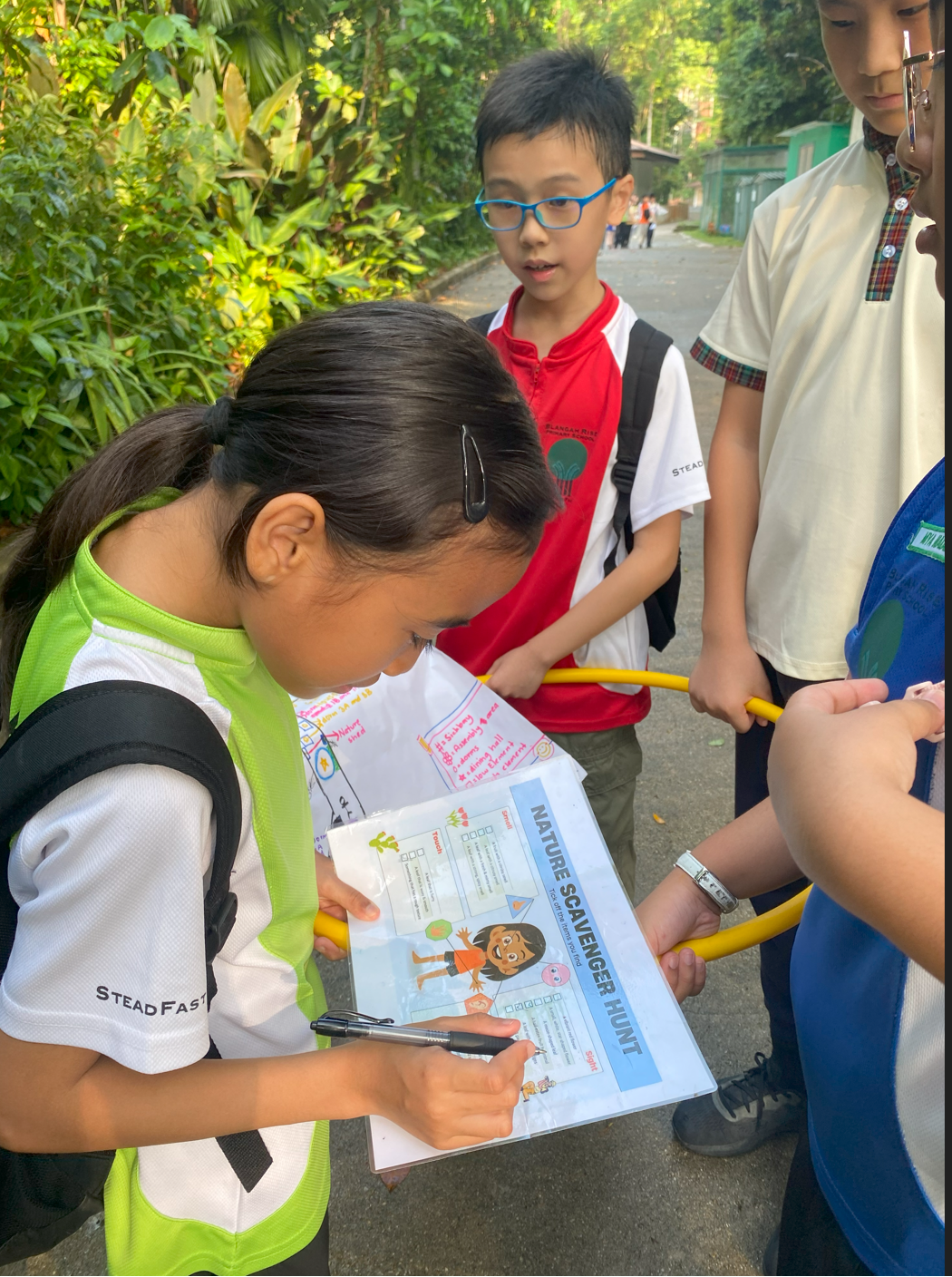 A girl writes on a Nature Scavenger Hunt worksheet while three other children stand nearby outdoors.