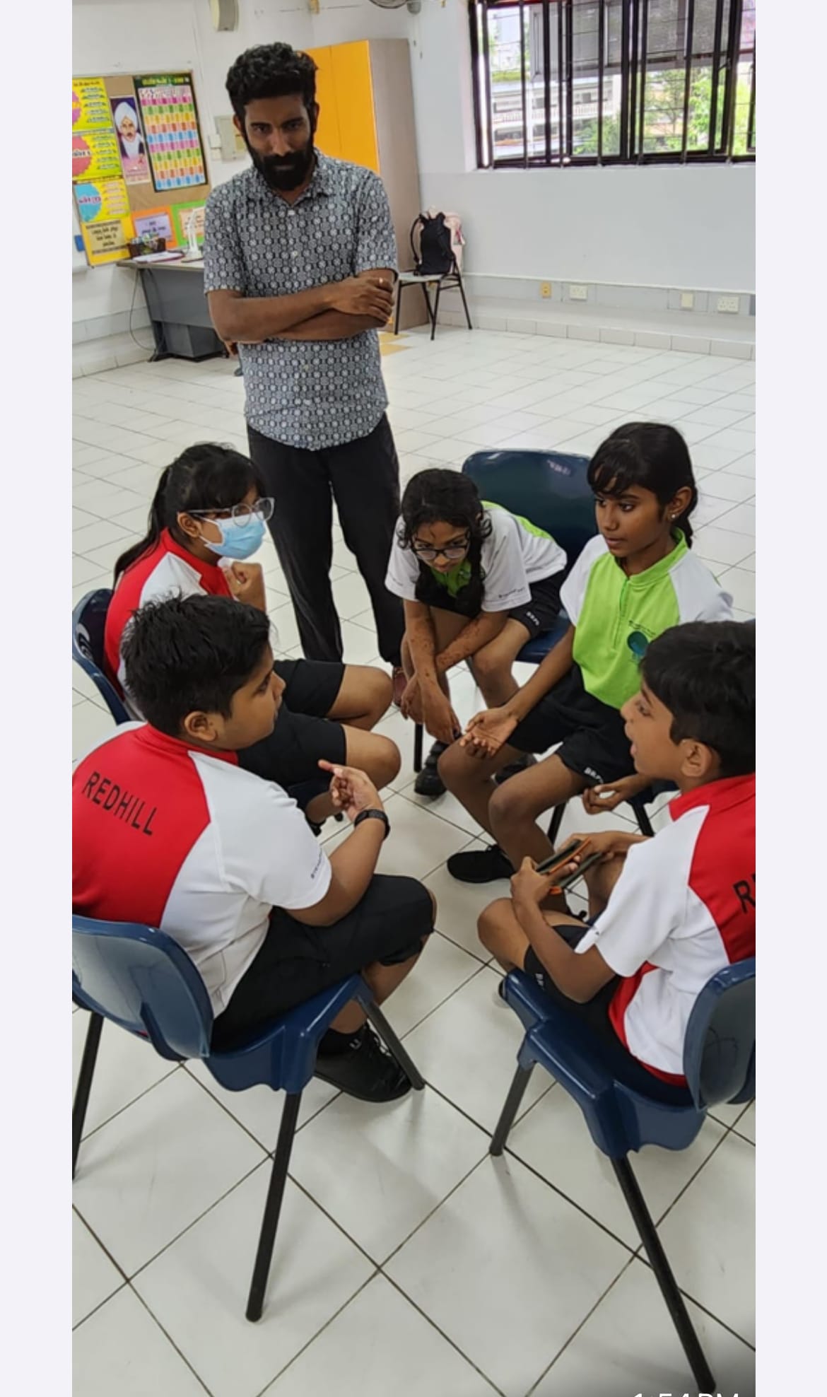 A teacher with crossed arms stands watching five students in Redhill uniforms seated in a circle discussing indoors.