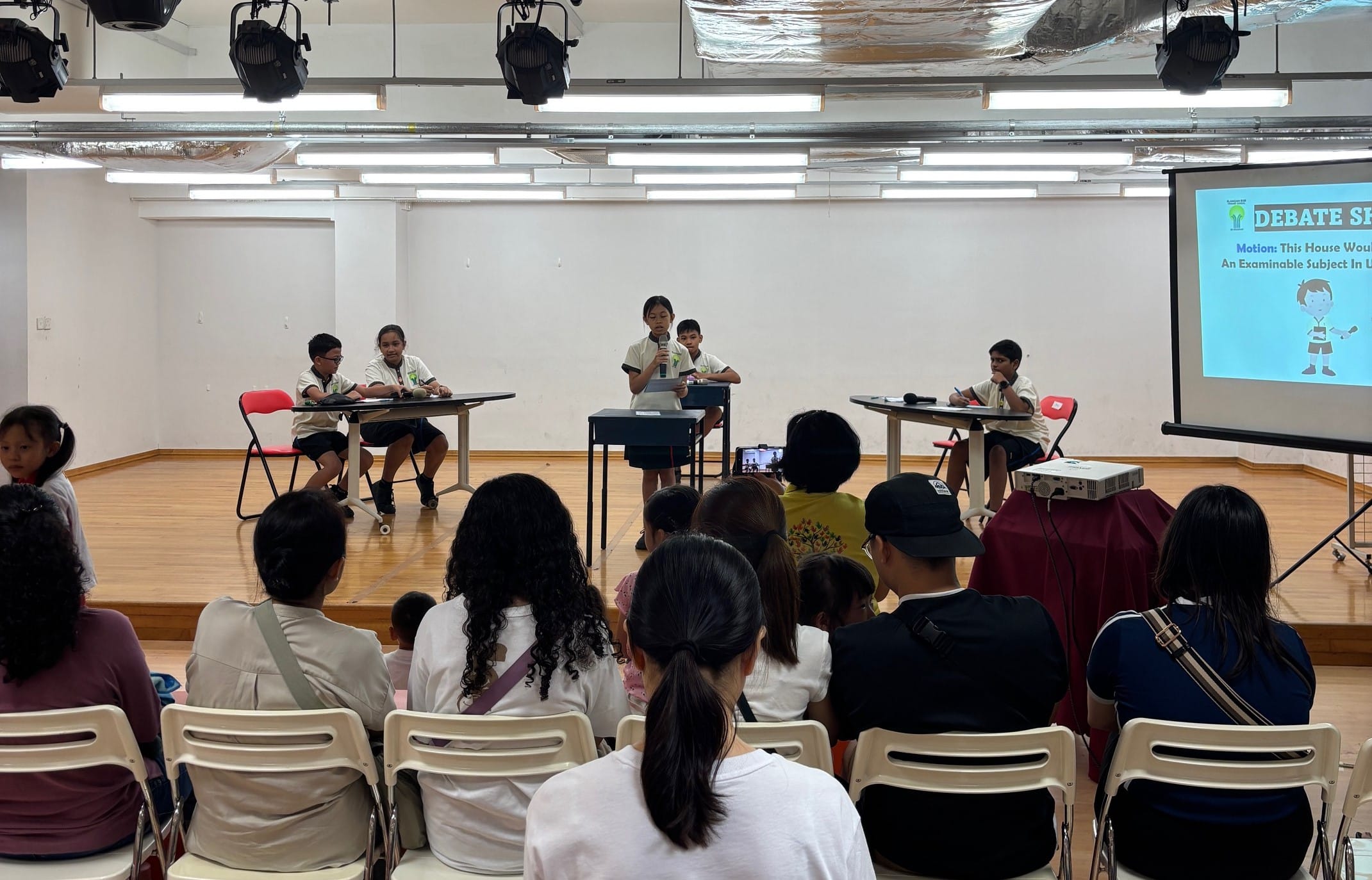 A student speaks into a microphone at a school debate event while an audience watches and a projection screen displays "Debate."
