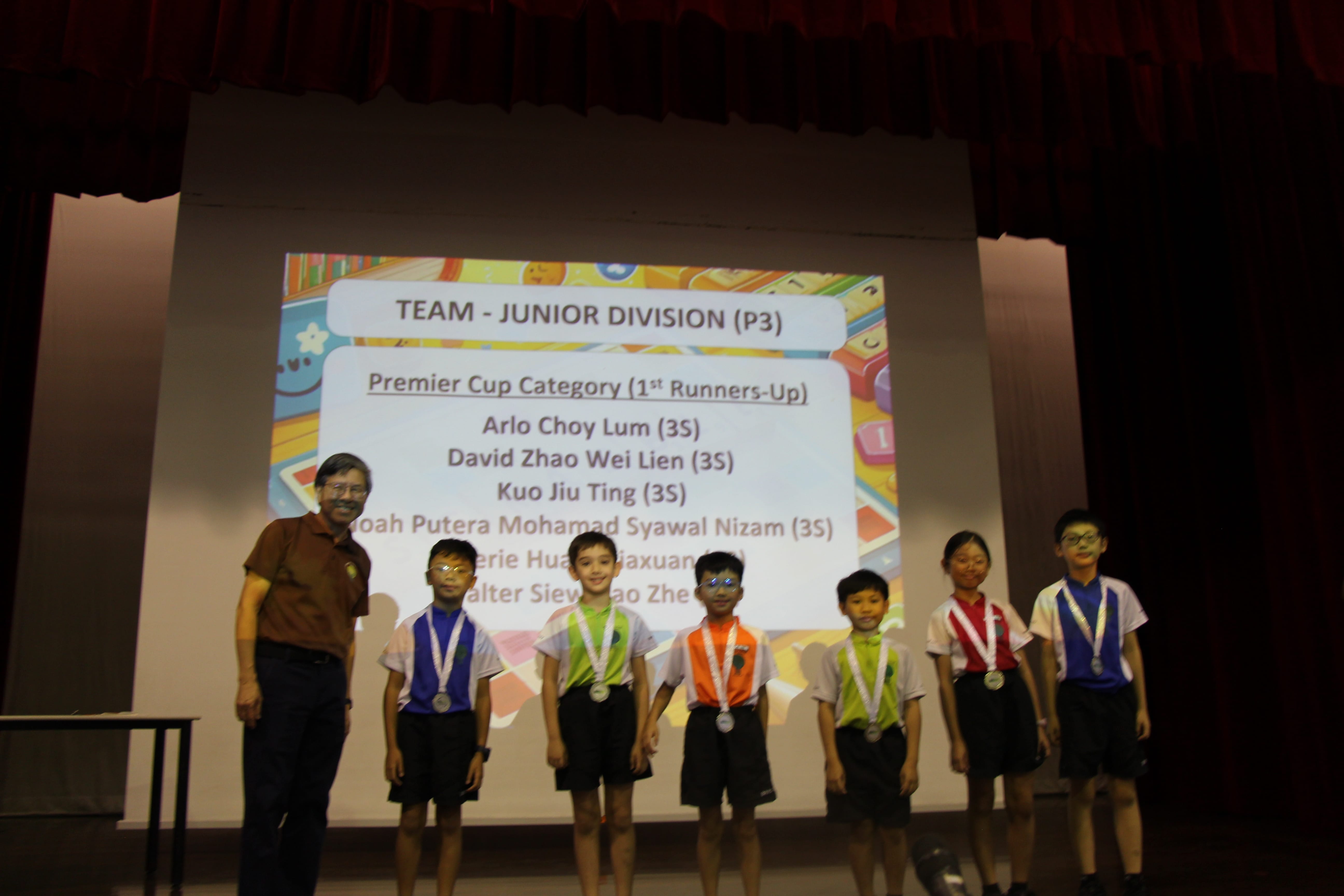 Teacher and six students with medals pose on stage in front of projected competition results.