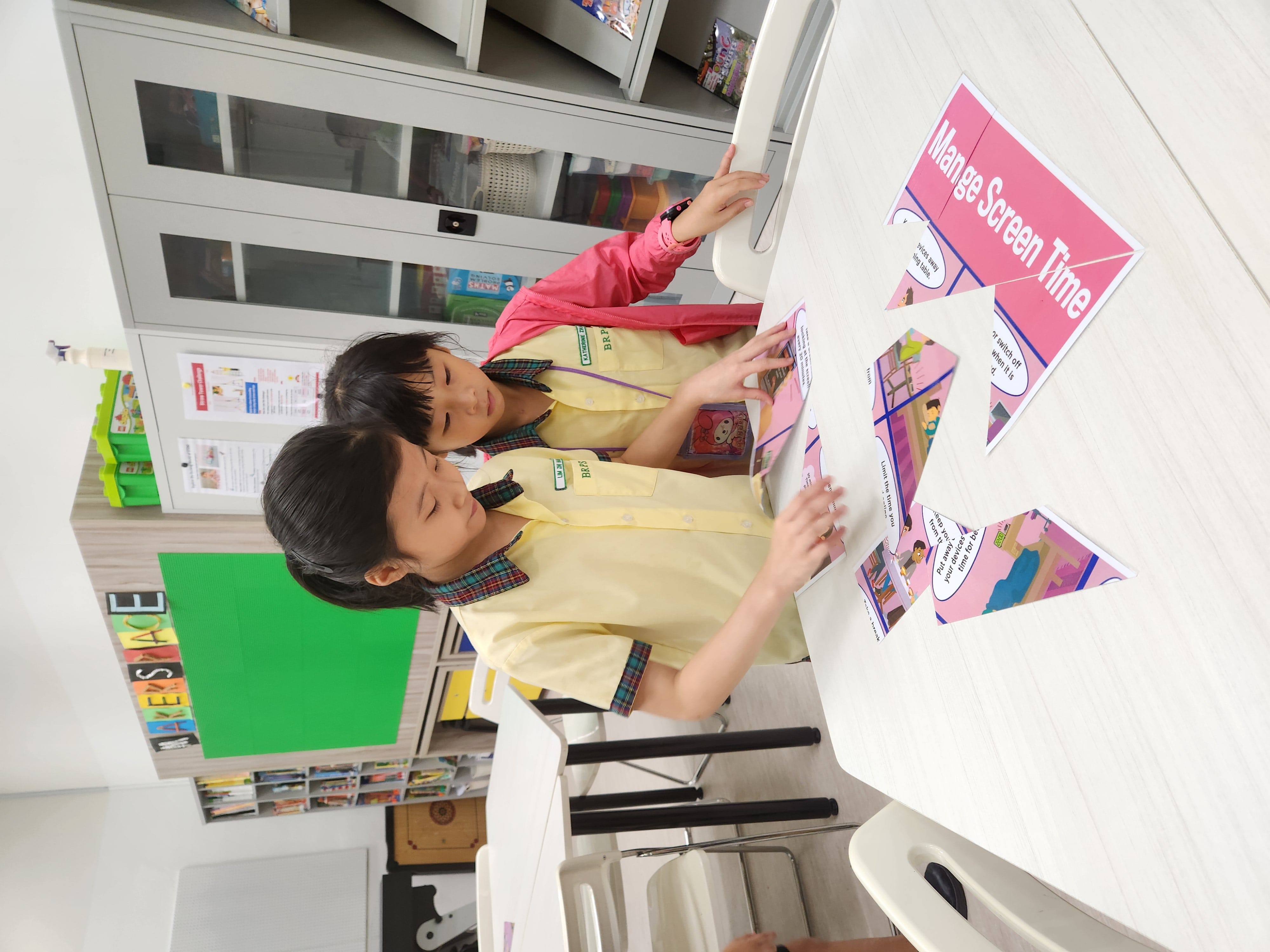 Two young girls in school uniforms arrange pink comic strip cards about managing screen time on a white table.