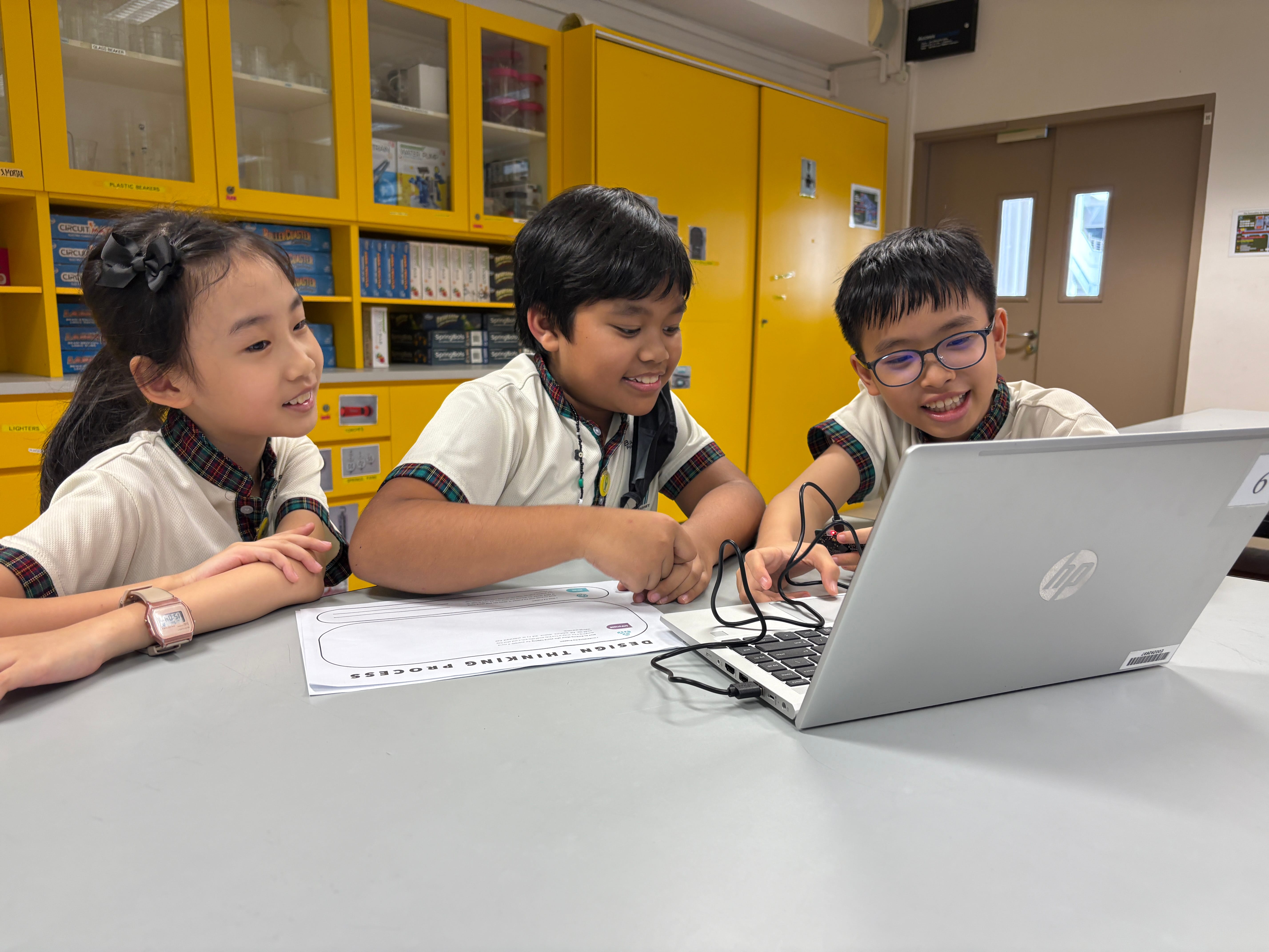 Three smiling school children in uniforms collaborate on an HP laptop at a desk with a design thinking worksheet.