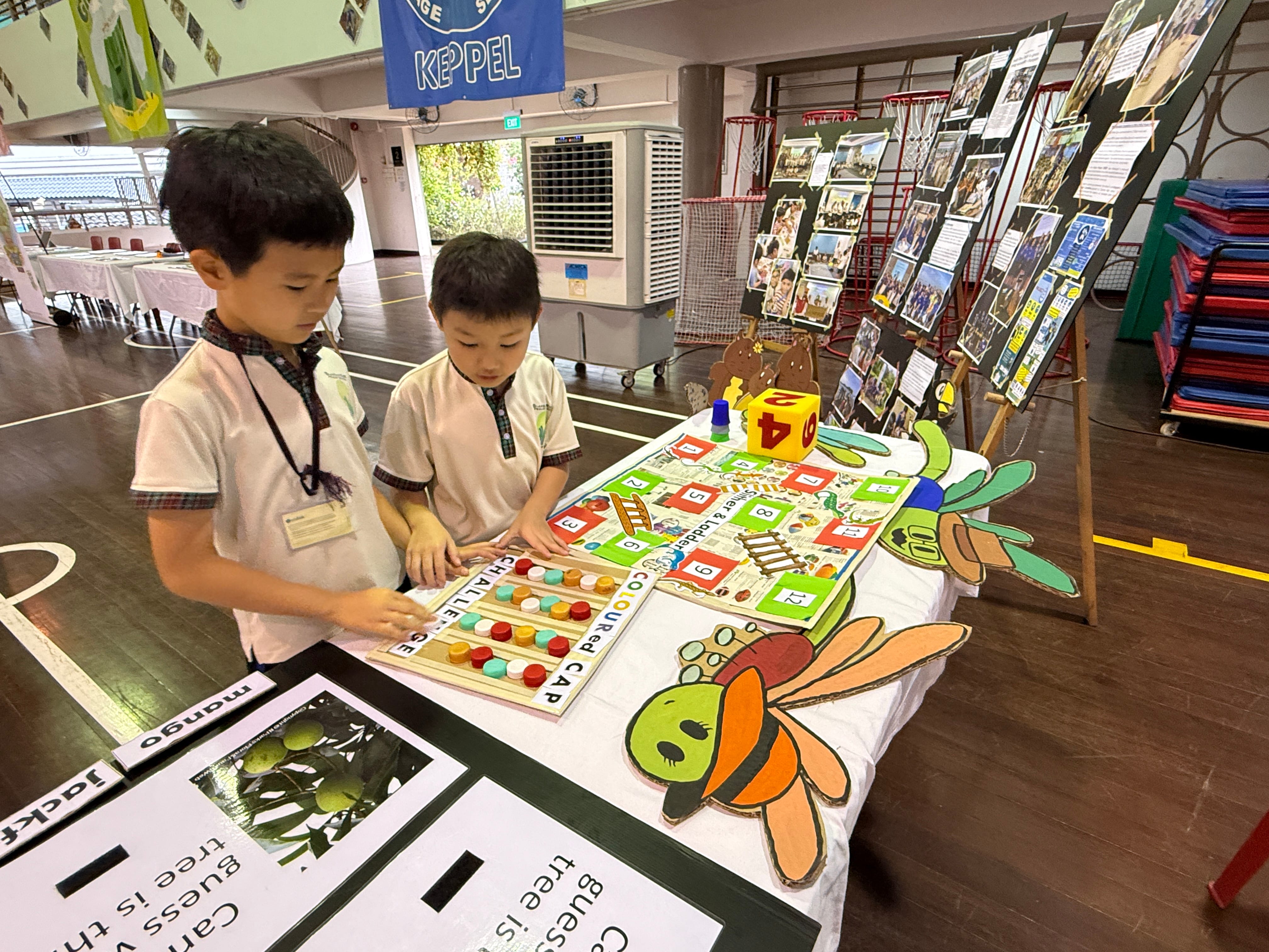 Two young boys in school uniforms play a colorful snakes and ladders board game at a table during a school event.