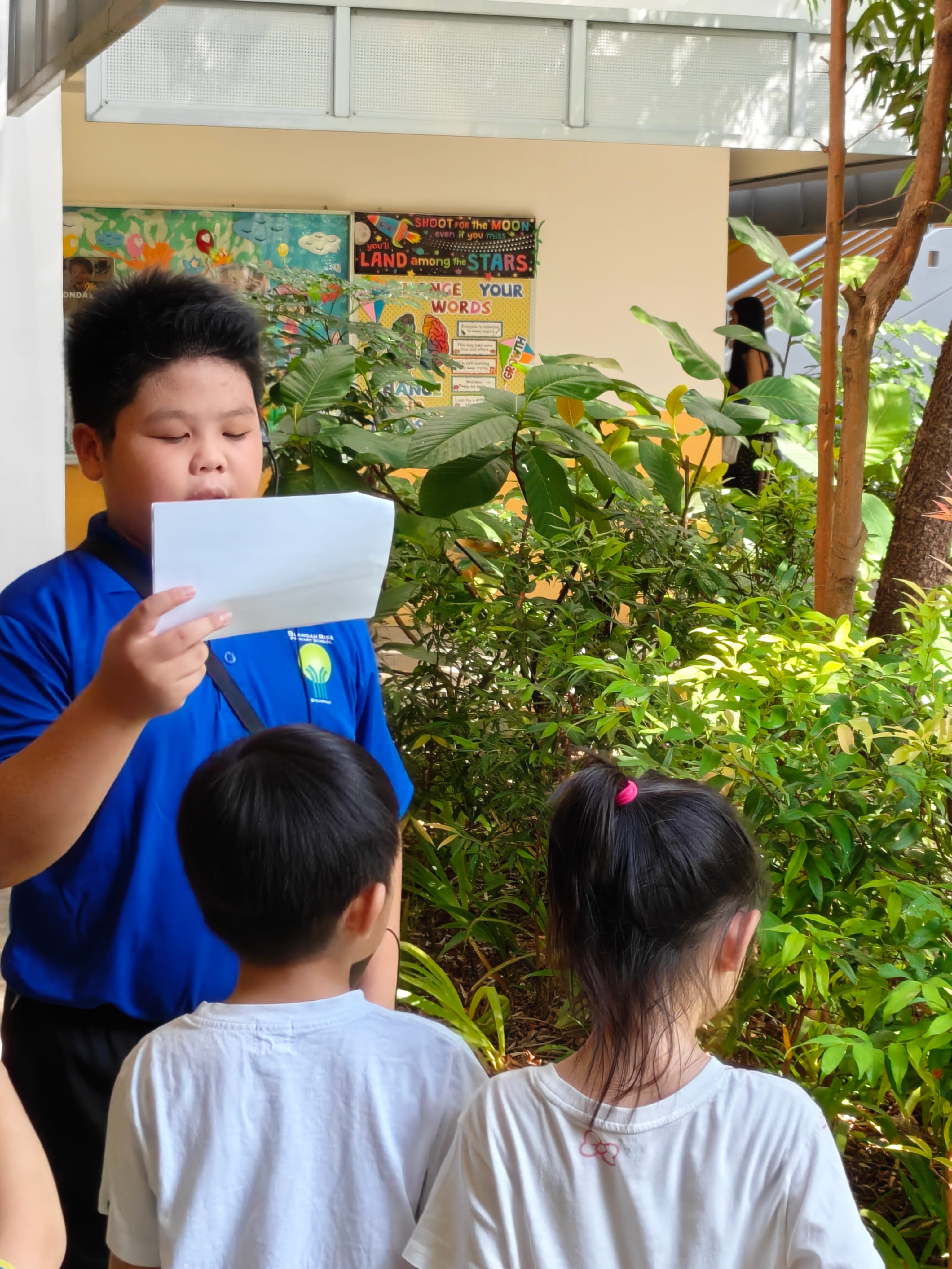 A boy in a blue shirt reads from a paper to two younger children outdoors near a school wall with motivational posters.