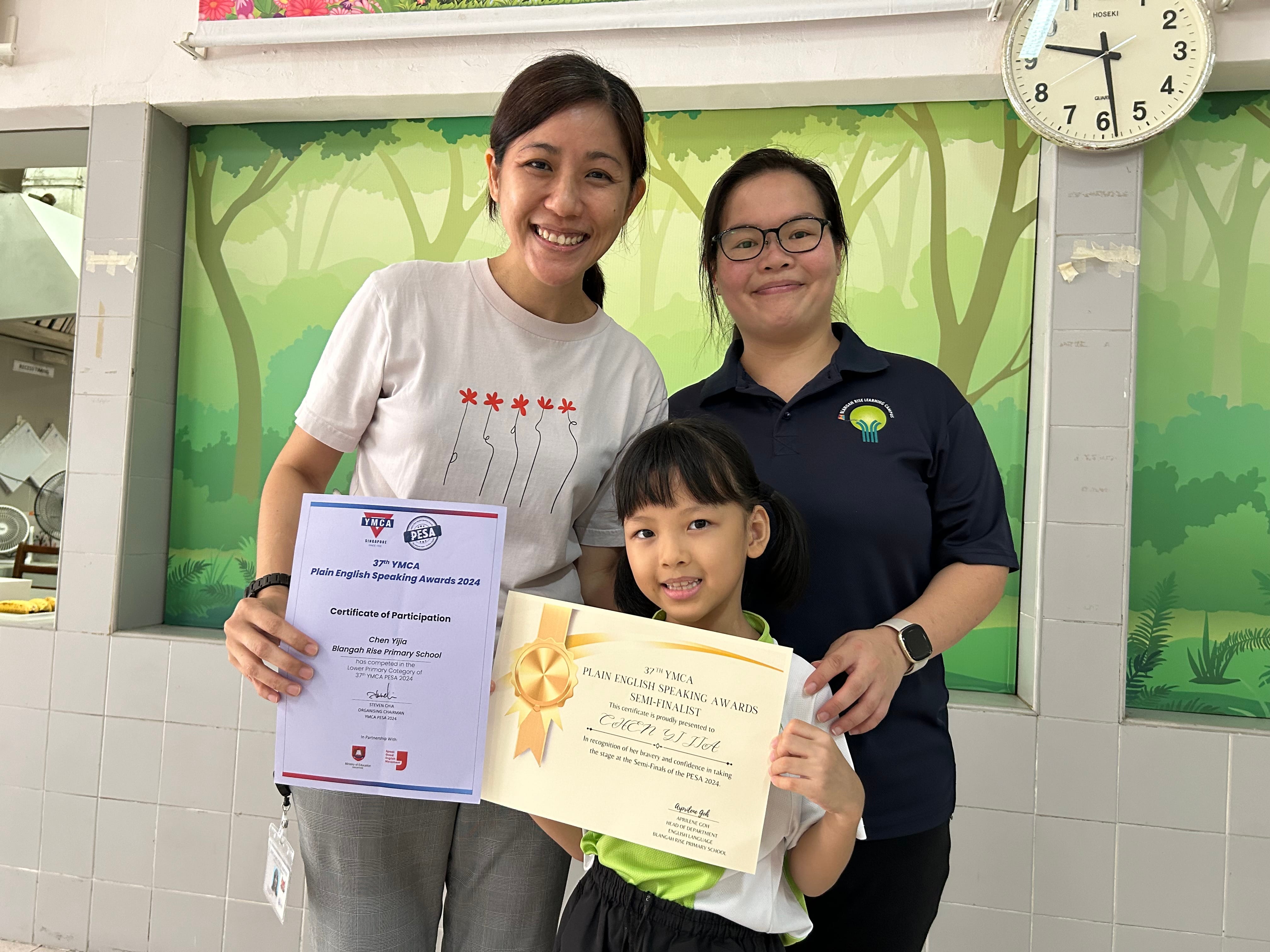 Two women and a young girl pose holding YMCA Plain English Speaking Awards 2024 certificates indoors.